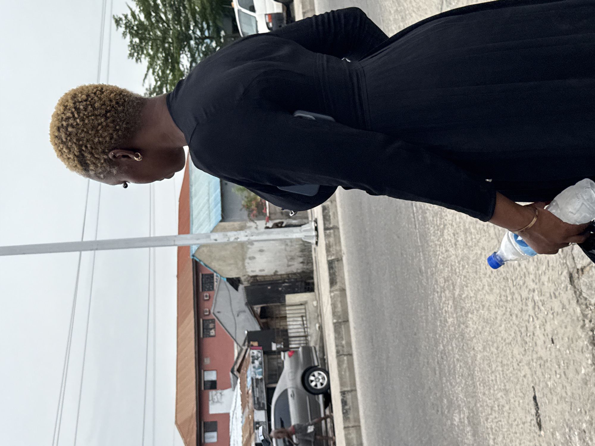 A woman with short, blonde, curly hair, wearing a black dress with a belt, is walking on a city street. She is holding a plastic water bottle in her right hand. The street is adjacent to a building with a red roof. A gray car is parked on the street. Overhead utility lines are visible. The photo appears to be taken on a cloudy day in Ado, Nigeria.