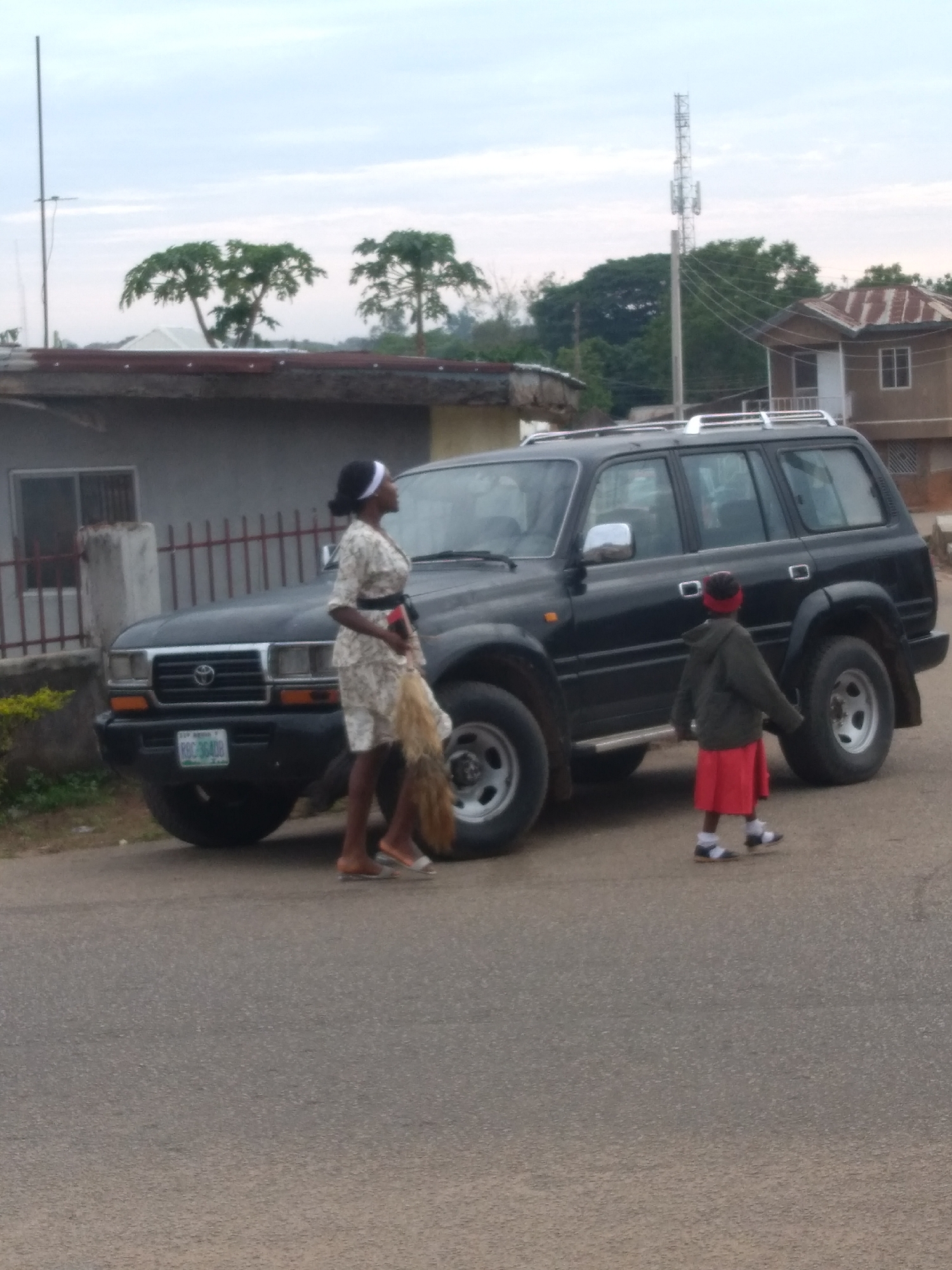 The image depicts a street scene in Jos, Plateau State, Nigeria, identified by the "PLATEAU" inscription on the vehicle's license plate. A dark-colored, four-door Toyota Land Cruiser SUV, model J80 series, is positioned diagonally on the right side of a paved road. The vehicle displays license plate "RYG-364UD" on its front bumper. It is equipped with silver roof rails.
A dark-skinned adult female stands to the left of the SUV, facing right. She wears a white headscarf, a light-colored patterned short-sleeved dress with a dark belt, and light-colored sandals. Her right arm extends down, holding a light brown, fibrous object resembling a traditional broom or whisk, which rests against her hip. Her gaze is directed upwards and towards the right.
A small child is located to the right rear of the SUV, walking towards the right side of the frame with their back partially to the camera. The child wears a dark green long-sleeved jacket, a bright red skirt, white socks, and dark sandals. A red band is visible on the child's head.
The background includes several structures and natural elements. To the left, a light-colored building with a red corrugated metal roof is partially visible, featuring a window with bars and a fence with dark red vertical railings. Behind this building, several trees with large, palmate leaves (possibly papaya trees) are prominent against the sky. To the right, a brown building with a distinct reddish-brown, multi-peaked roof and a white window frame is present. Further in the background, a tall, grey communication tower with multiple antennae rises above other trees. The sky is overcast with a light grey-blue hue. A slender, tall metal pole is visible on the far left edge of the image. The road surface is dark grey asphalt.
