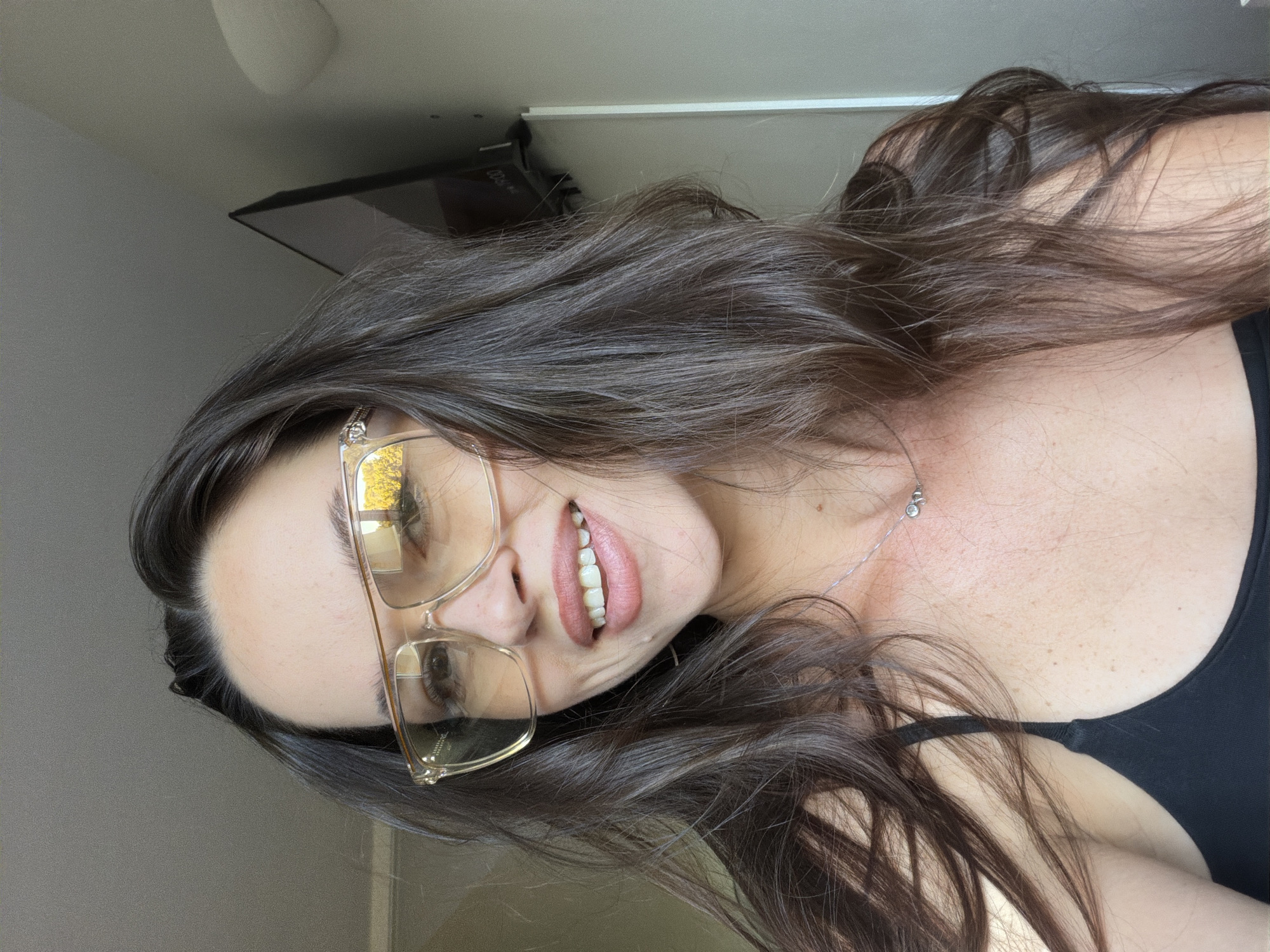 A young woman with long, dark brown hair is looking directly at the camera, smiling. She is wearing oversized, clear-rimmed glasses with yellow-tinted lenses. Her lips are painted a nude pink color, and she has a delicate silver necklace with a small charm around her neck. She is wearing a black tank top. The background appears to be an indoor setting with natural light filtering in, possibly a room in Salou, Spain, given the context. A television screen is visible on the wall in the background, and the overall lighting suggests it is daytime.