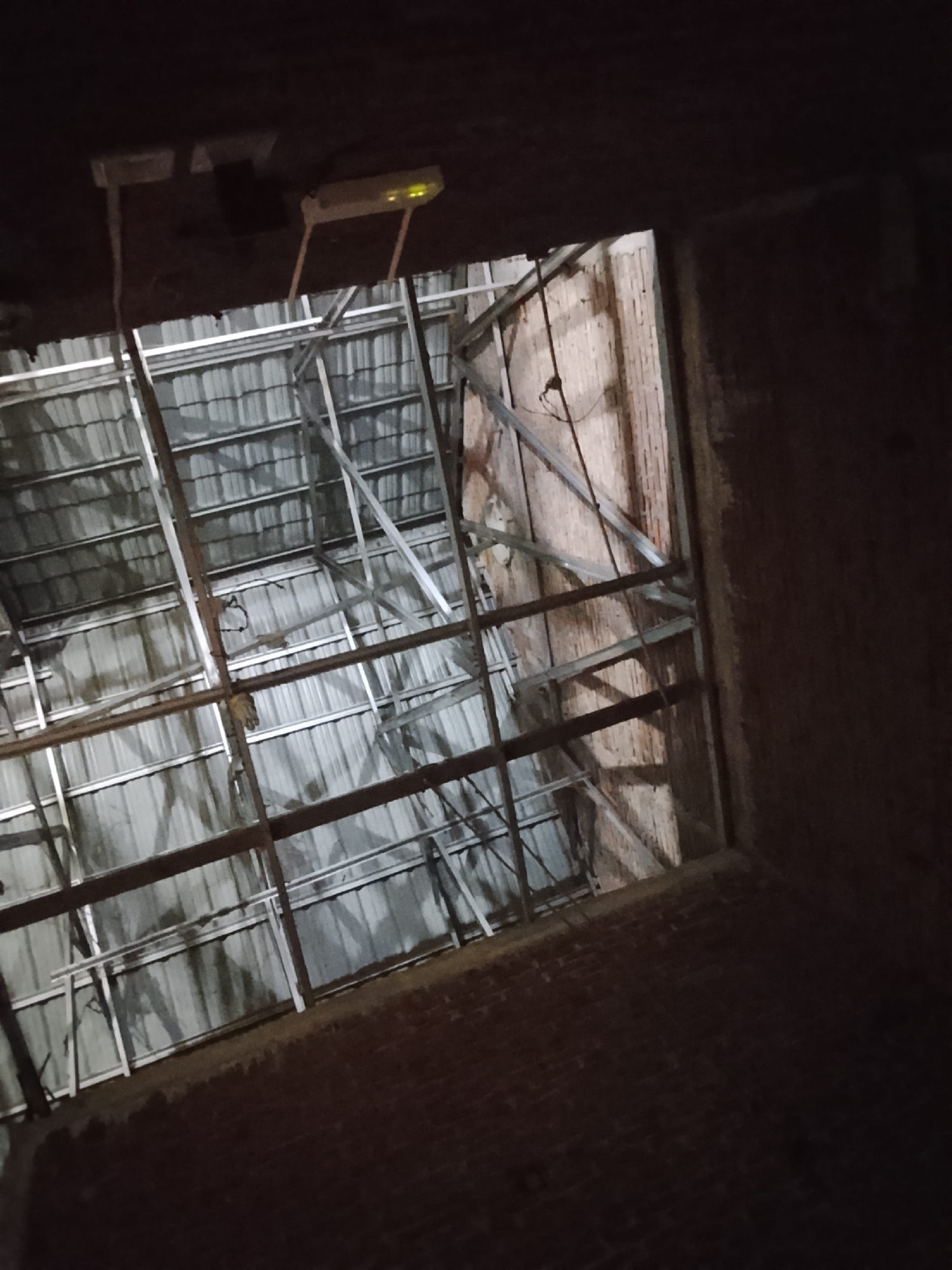 The image shows a view looking up into an unfinished or abandoned building. The ceiling is composed of corrugated metal sheeting supported by metal framing. Some brickwork is visible on the right side, indicating a wall. In the upper portion of the image, there are some electrical conduits and a device that appears to be a Wi-Fi router or a similar network device mounted on the ceiling. The lighting is dim, suggesting it might be nighttime or an interior space with limited illumination. There are no people or animals visible in the frame. The overall impression is one of disrepair or ongoing construction. The specific location in Tanjongbabahkrueng, Indonesia, cannot be determined from the visual information alone.