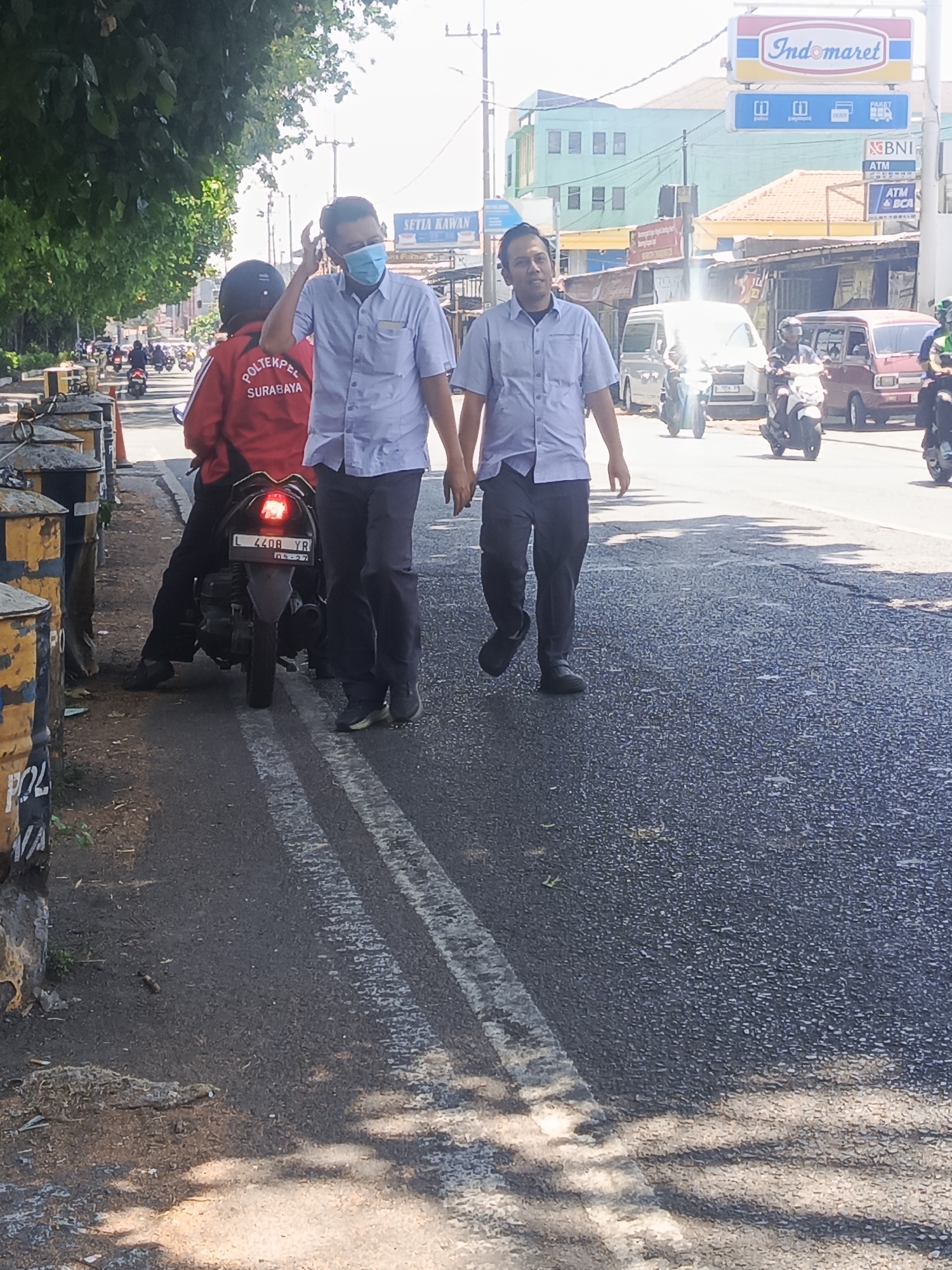 In Kedungrejo, Indonesia, two men in light blue collared shirts and dark pants are walking on an asphalt road. The man on the left is wearing a light blue face mask and has his right hand raised to his ear. He is walking towards the right of the frame. The man on the right is looking towards the viewer and has his right hand resting on his left arm. They are walking in the same direction. Behind them, a person on a red motorcycle with "POLTEKPEL SURABAYA" written on the back is seated. The motorcycle license plate reads "L 4408 YR". The environment is a street with trees on the left side, casting shadows on the road. In the background, buildings and signs are visible, including an "Indomaret" sign. Several other motorcycles are on the road, some with riders. The scene appears to be during the daytime, with bright sunlight.