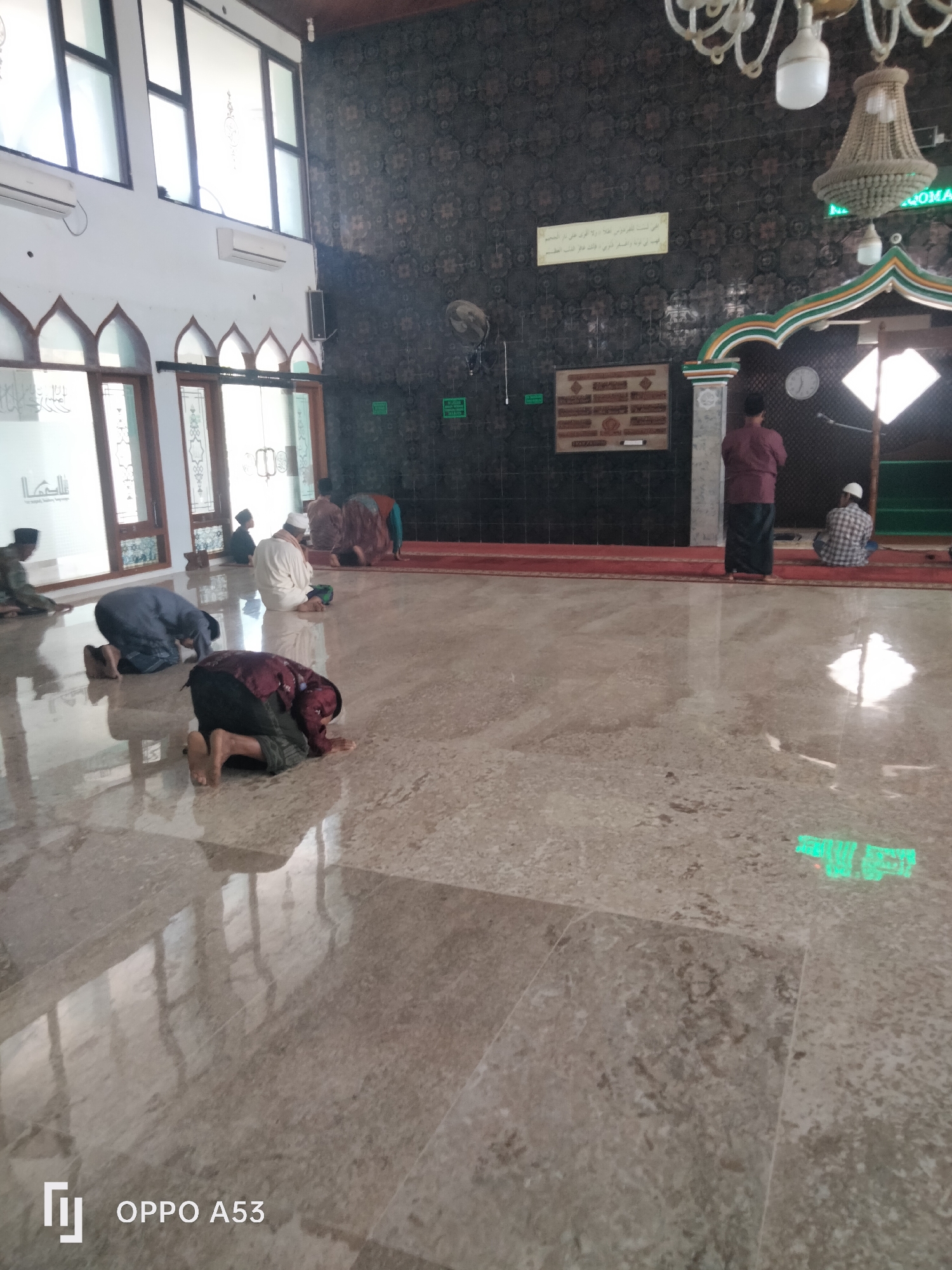 The image shows the interior of a mosque in Badung, Indonesia. Several individuals are engaged in prayer on the polished floor, which reflects the ambient light. The individuals are dressed in traditional Muslim attire, including kufis, sarongs, and shirts. The mosque features arched windows with decorative glass and air conditioning units mounted on the walls. The wall behind the prayer area is adorned with intricate patterns and calligraphy. A chandelier hangs from the ceiling, and an Islamic plaque is mounted on the wall. The floor is a light-colored marble with reflective qualities, showing the reflections of the ceiling and chandeliers. A prominent green sign with text is visible on the floor in the lower right section of the image, along with the text "OPPO A53" and a logo in the bottom left corner.