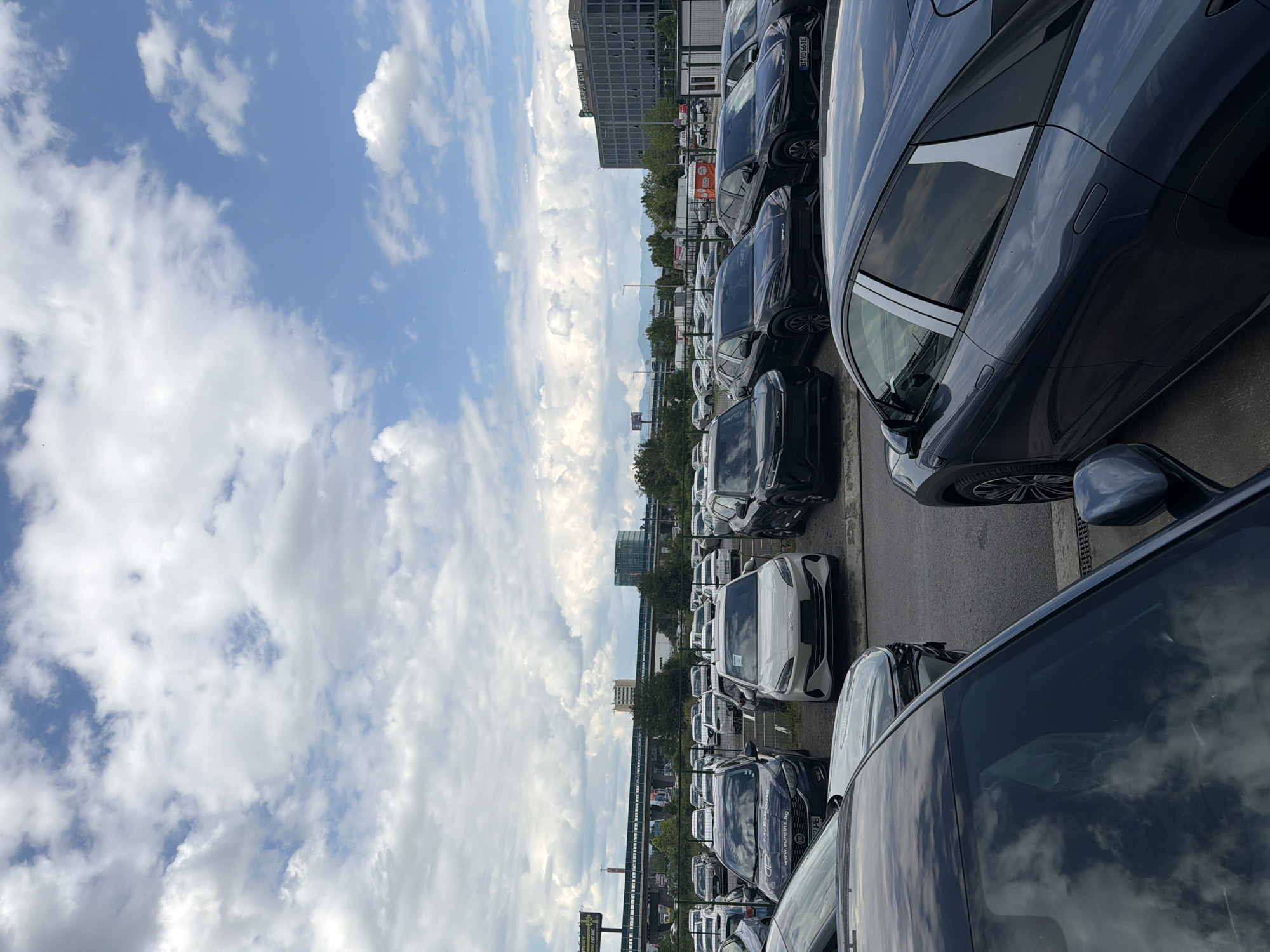 This image captures a wide outdoor car parking lot, likely a dealership or storage facility, under a bright blue sky filled with scattered white cumulus clouds. The perspective is slightly elevated, looking across numerous rows of neatly parked vehicles.
The majority of the cars are modern models, predominantly in dark colors such as black, dark grey, and dark blue, though some white cars are also visible. Reflections of the sky and clouds are prominent on the car bodies and windows. Several Bulgarian license plates are visible, including "B 9VD688E" and "02 PB", indicating registration in Sofia. One car displays a sticker with the website "www.eurons.bg".
In the background, a large, multi-story building with dark windows stands out. Text on the building identifies it as "EURO SPORT CENTER" and "LEXC". Beyond the buildings, distant mountains are faintly visible, contributing to the suburban or semi-urban setting. The scene is illuminated by clear daylight, suggesting a sunny day. No individuals or specific activities are depicted; the image provides a static view of the car lot in Busmantsi, Bulgaria.