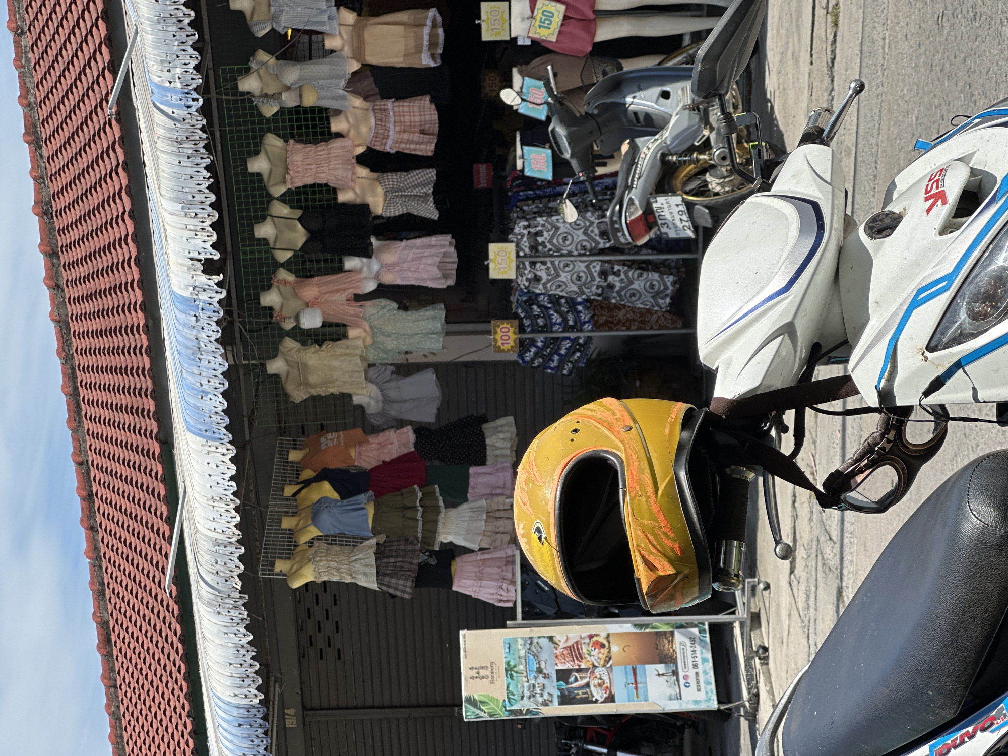 A vertically oriented medium shot shows a roadside shopping area in ตำบลเกาะพะงัน, Thailand, cluttered with various items. A red-tiled roof overhangs the storefront, under which clothes displayed on mannequins line the shop. Price tags of 150 baht are visible on some garments. A pair of motorcycles are parked in front of the store. A yellow helmet rests on one of the motorcycles. A sign promoting "Harmony" is nearby, with a phone number for reservations: 061-514-2452. The license plate of one of the motorcycles is partially visible and reads "3 กญ กรุงเทพมหานคร 2791".