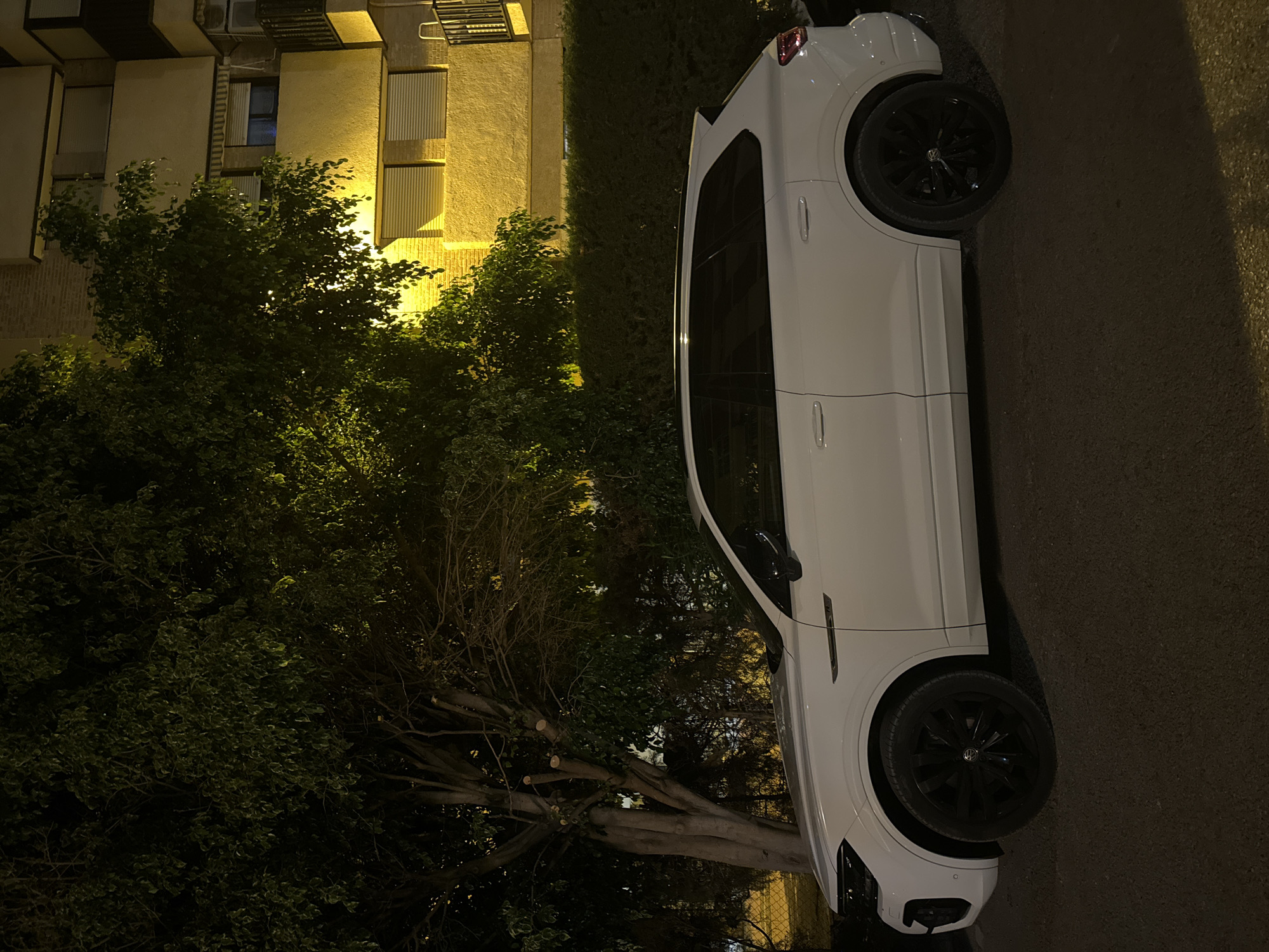 A white Volkswagen Touareg is parked on an asphalt surface in front of a building at night. The car is positioned in profile, facing left. The building behind the car is illuminated by yellow lights, revealing its yellow facade and several windows. To the left of the car, there is a large tree with dark green leaves and a thick trunk. The overall lighting suggests it is nighttime. The scene appears to be an urban setting, possibly in the city of Murcia, Spain, given the architectural style of the building and the type of vegetation. No people are visible in the image.