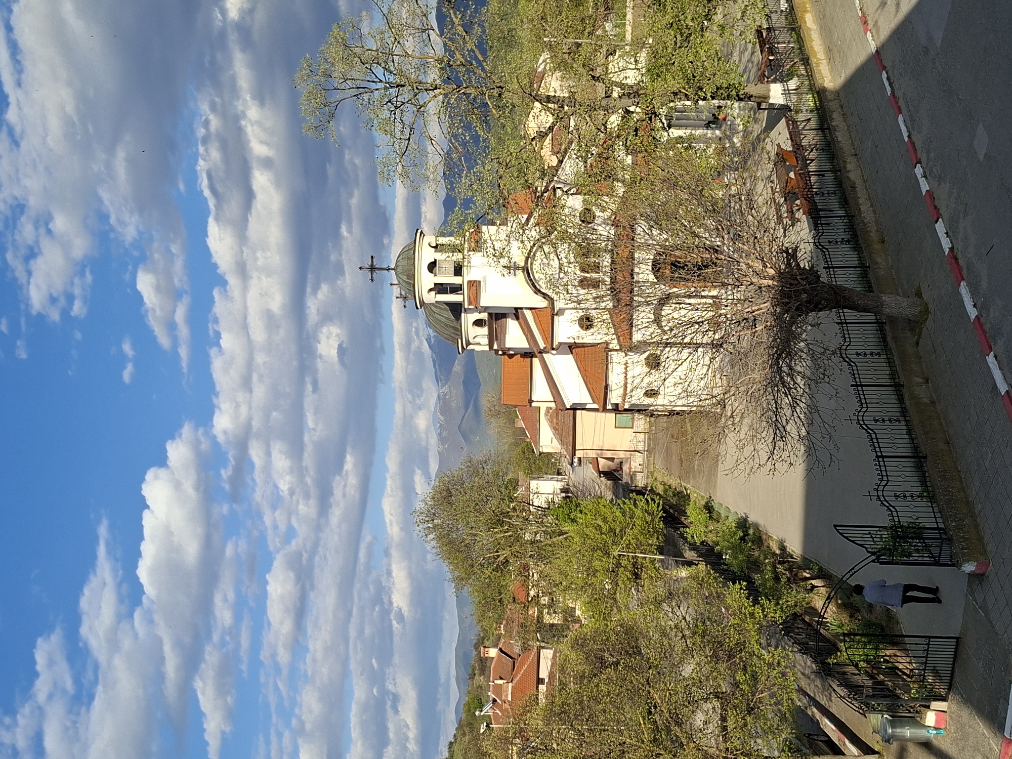 The image shows a view of a white, two-story building with a cross on top, likely a church, in an urban area. The sky is blue with scattered clouds. Buildings with red tile roofs are visible among green trees. There is a street with a black iron fence and a person walking near a gate. The location is in the city of Unknown, Unknown.