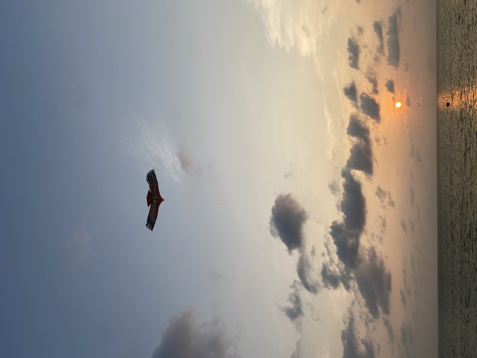 The image depicts a sunset scene with a kite flying in the sky. The location appears to be Choeng Thale, Thailand, based on the coastal aspect. The kite, shaped like a bird, is positioned on the left side of the frame against a predominantly blue sky. A kite string extends from the kite toward the right side of the image. On the right side, the sky is filled with clouds, ranging in color from white to grey, with a visible setting sun. The setting sun is reflected in the calm ocean water. The overall scene has a tranquil atmosphere.