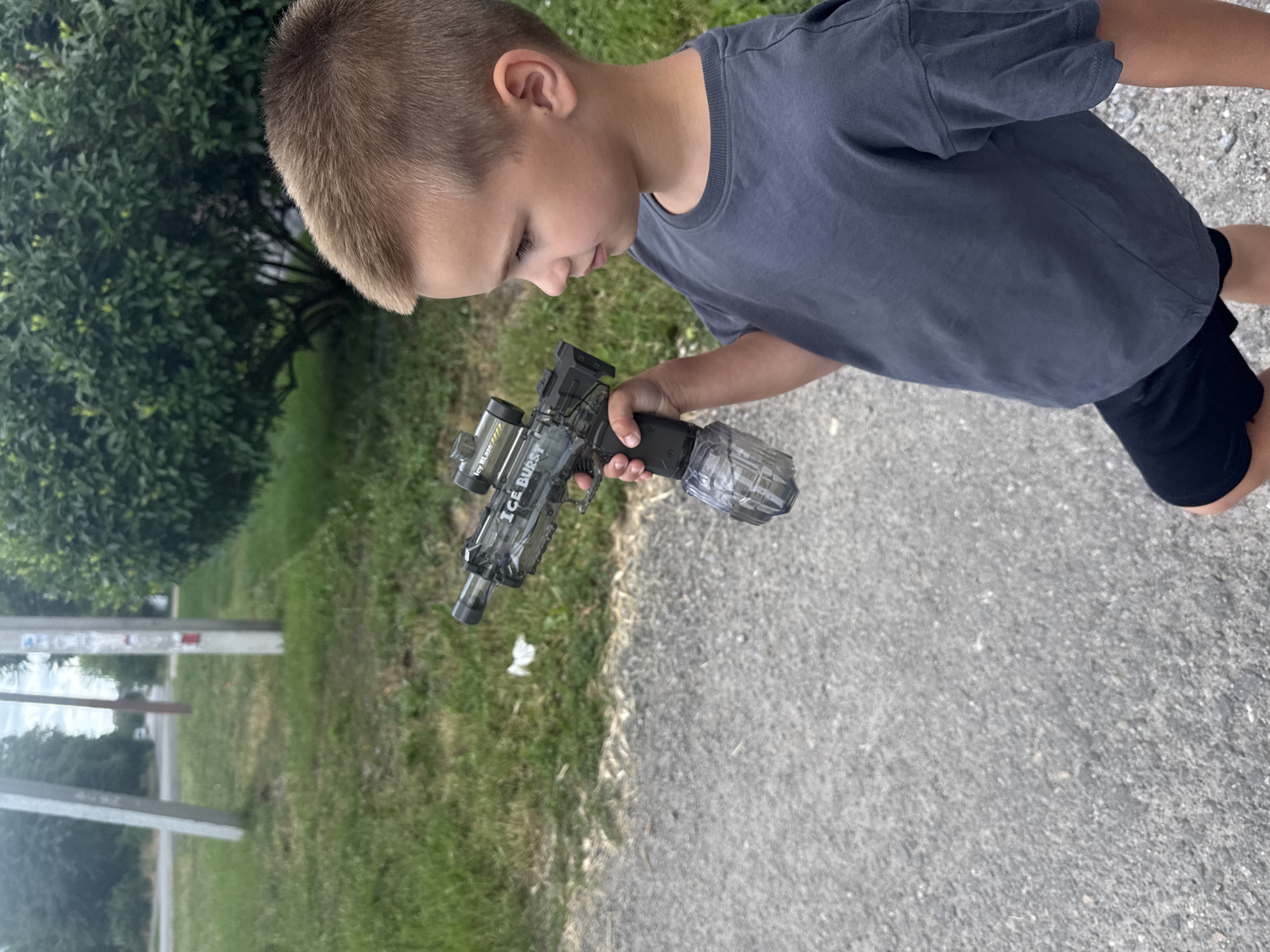 A young boy stands outdoors, holding a toy gun. The boy is wearing a dark grey t-shirt and black shorts. The toy gun is translucent and has a drum magazine attached. He is looking at the toy gun. The background consists of a grassy area, trees, a paved surface, and some utility poles. The lighting suggests it is daytime. The scene appears to be in a suburban or residential area.