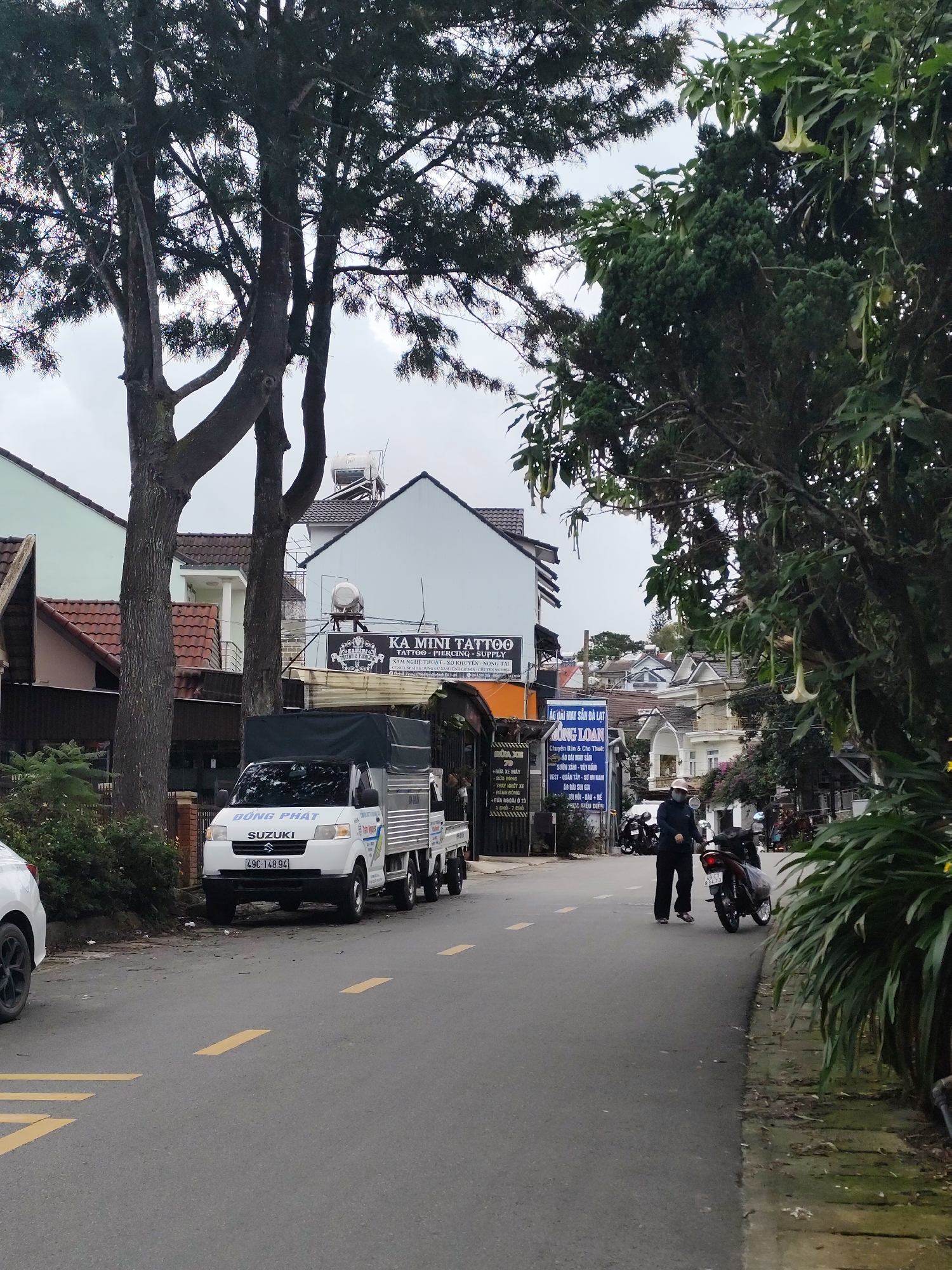The image depicts a street scene in Da Lat, Vietnam. A white Suzuki van with the license plate "49C-148.94" is parked on the right side of the road. A person wearing dark clothing and a hat is walking away from a motorcycle on the left side of the road. The street is paved with asphalt and has yellow painted lane dividers. Buildings line the street, including a structure with a sign that reads "KA MINI TATTOO." Lush green trees with hanging flowers are visible on both sides of the street. The sky is overcast, suggesting a cloudy day.