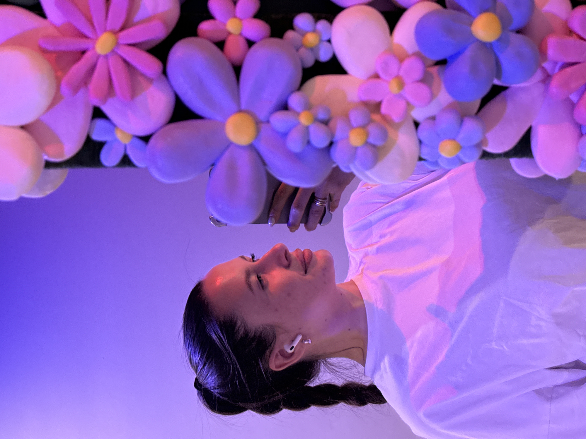 The image, presented rotated 90 degrees counter-clockwise, features a young woman looking upwards, surrounded by large, colorful decorative flowers. In its natural orientation, the woman is seen from a low angle, her head and upper torso filling the lower half of the frame. She has dark hair styled in a braid and wears a white top. A white wireless earbud is visible in her right ear. Her right hand, adorned with a ring, holds a smartphone, which reflects the vibrant colors of the flowers above.
The prominent flowers, likely soft sculptures or balloons, are in shades of pink, purple, and blue, each with a bright yellow center. They are arranged overhead, creating a whimsical and immersive environment. The background is a solid gradient of purple and lavender, indicating an artificially lit, indoor setting.
The woman's expression is calm and engaged, suggesting enjoyment as she looks at the artistic display, possibly interacting with it by taking photos or videos with her phone. The scene appears to be part of an art installation, a themed event, or a photo opportunity area. There is no visible text in the image. The location context for this scene is Vniissok, Russia.