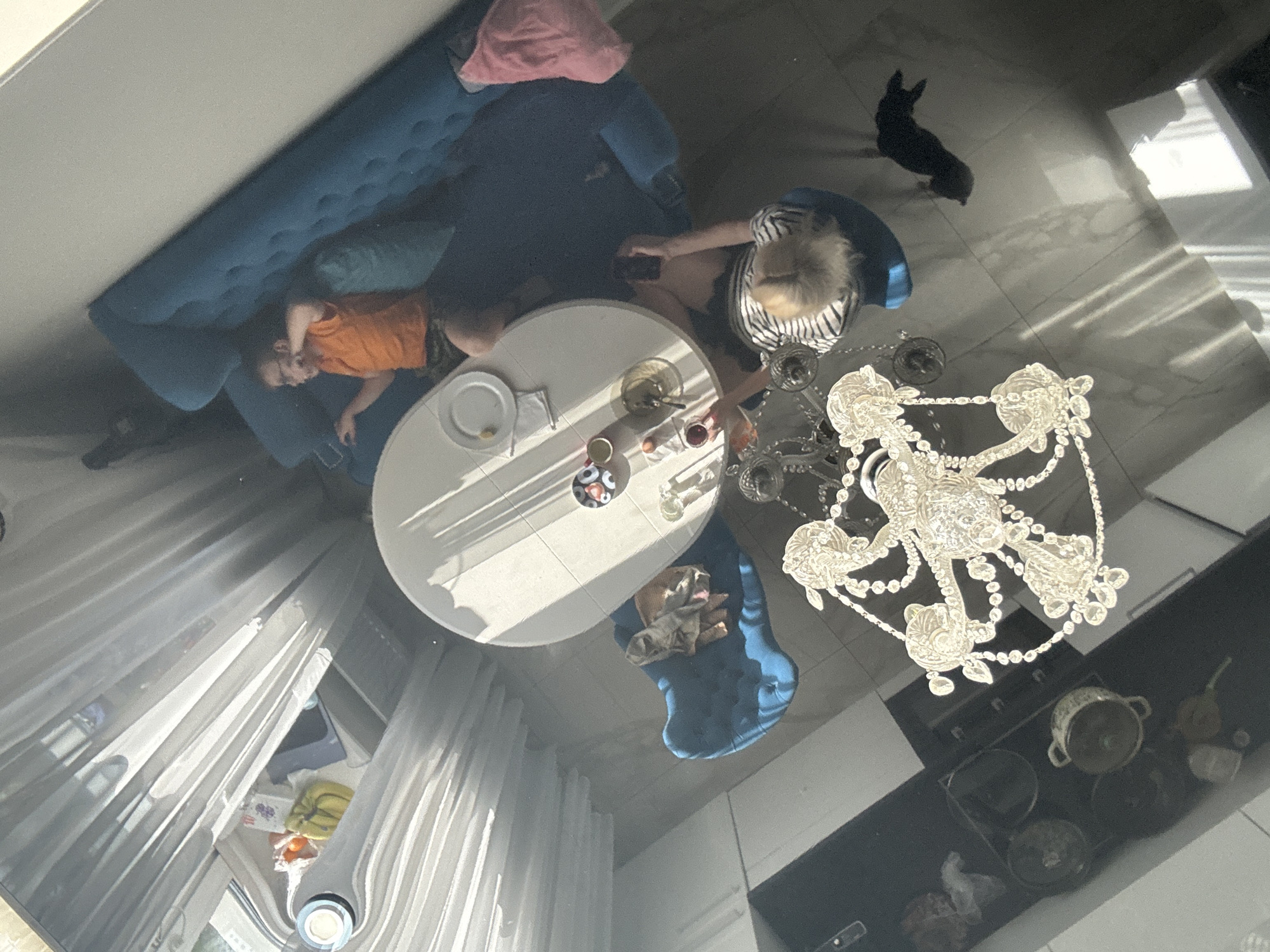 This image depicts a high-angle, possibly reflected, view of a living room or dining area. The primary subjects are a young boy and a woman, along with a small black dog and a chandelier.
The boy, wearing an orange t-shirt and patterned shorts, is lying down on a blue sofa, appearing relaxed or asleep. A pink blanket is draped over a portion of the sofa. The woman, with blonde hair and wearing a striped top and dark shorts, is seated in a chair near a round white table. She is holding a mobile phone.
On the table, there are breakfast items, including a plate with a napkin and what appears to be a piece of fruit, a bowl with some food, an egg, and a glass of red liquid, possibly juice or wine. The chandelier above the table is ornate and made of crystal, reflecting light. The dog is seen standing on a tiled floor in the background, casting a shadow.
The scene is illuminated by sunlight streaming through sheer curtains, creating prominent light patterns on the floor and furniture. This suggests it is daytime, likely morning or afternoon. The overall atmosphere appears calm and domestic. The presence of curtains and what looks like a window area to the left suggests an indoor setting. The floor is tiled with a marble-like pattern. In the background on a dark surface, possibly a countertop or cabinet, are various kitchen items, including pots.
No visible text is present in the image.