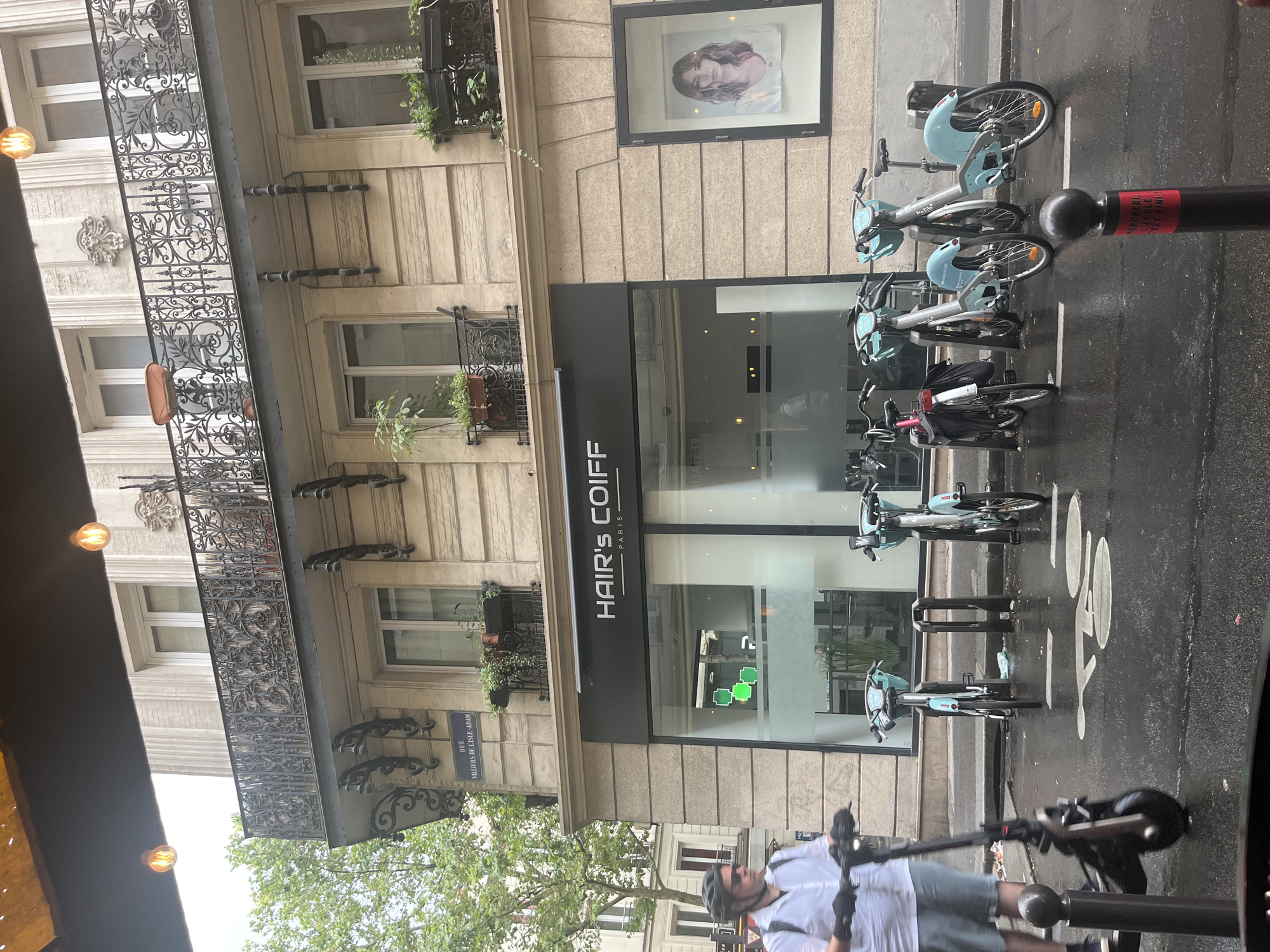This image captures a street scene in Paris, France. The primary subjects are several public bicycles parked along the sidewalk, a storefront with the name "HAIR'S COIFF PARIS" visible, and a person on a scooter in the foreground. The bicycles are a light blue color and appear to be part of a bike-sharing program. The storefront is modern with a large glass window, and an advertisement featuring a woman's portrait is displayed higher up on the building.
The scene suggests an urban environment on a potentially wet day, as the asphalt appears dark and reflective, indicating recent rain or damp conditions. The weather is overcast, with no direct sunlight visible, contributing to a diffused lighting. The time of day is difficult to determine precisely, but the overall lighting suggests it is daytime.
Notable details include the ornate black ironwork of a balcony on the building, which adds a touch of classic Parisian architecture to the modern elements. The presence of street signage and the style of the buildings further confirm the Parisian setting. The person on the scooter is wearing a helmet and appears to be in motion, though their face is partially obscured. The overall atmosphere is one of everyday urban life, with transportation options like bicycles and scooters common in the city.
Visible text includes "HAIR'S COIFF PARIS" on the storefront and some unreadable text on a red and black pole next to the bicycles. There is also a street sign that reads "RUE VILLIERS DE L'ISLE-ADAM".