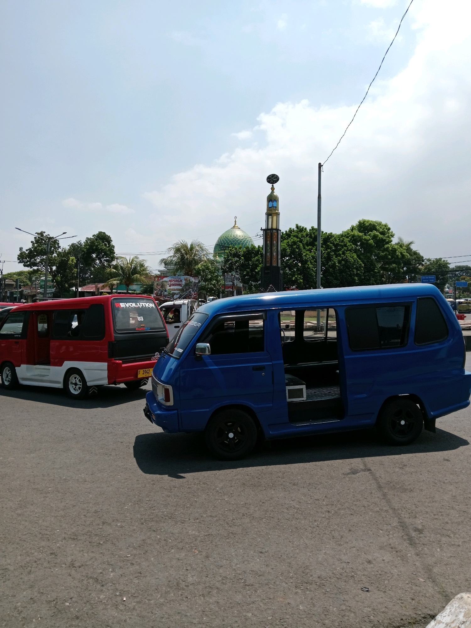 In Cianjur, Indonesia, under a partly cloudy sky, two vans are parked on an asphalt road. A bright blue van is in the foreground, its passenger side door open. To its left, a red and white van is visible, with the word "EVOLUTION" on its rear. In the background, a mosque with a green dome stands tall, flanked by trees. Traffic lights and power lines crisscross the sky. The scene appears to be an intersection or a street corner, with the vans possibly waiting for passengers or stopped at a light.