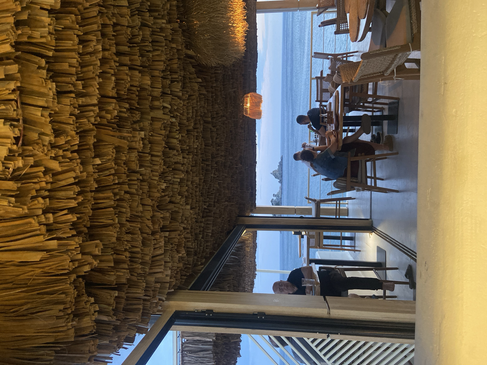 The image shows the interior of a restaurant in Petrovac centar, Montenegro, overlooking the sea. The ceiling is densely covered with a straw-like material. People are seated at tables, some dining and others looking at their phones. The restaurant has large windows providing a view of the coastline. A light fixture hangs above the dining area. One man is seated at a table close to the window with a beverage in front of him. The furniture appears to be primarily made of wood.