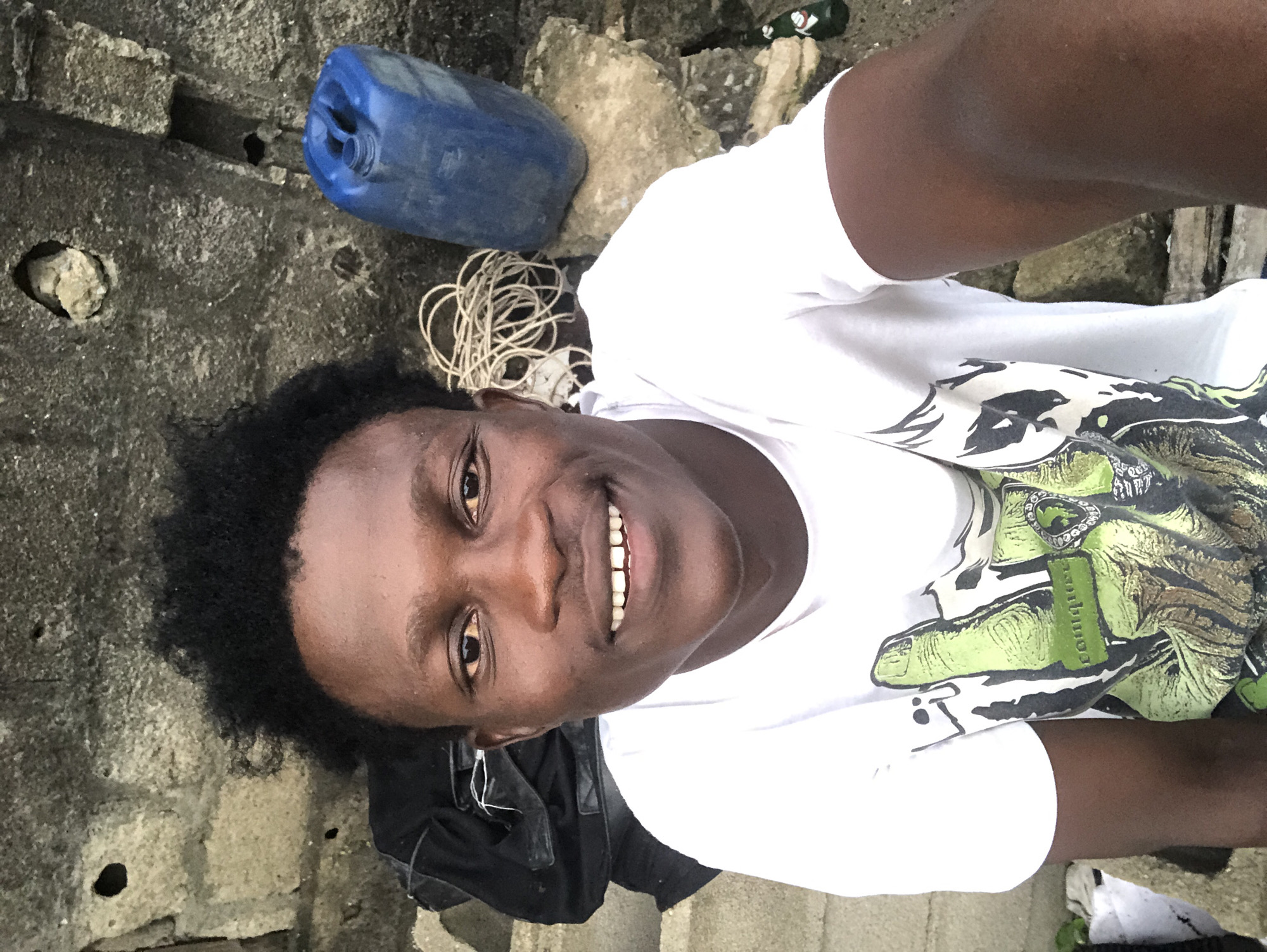 A young Black male is photographed from the chest up, looking directly at the camera with a wide smile. He has dark, curly hair and dark eyes. He is wearing a white t-shirt with a graphic design of a green hand. He is sitting outdoors with a rough, grey stone wall behind him. To his right, there is a blue plastic container with a lid and a coiled rope. To his left, a black bag is visible. The scene appears to be located in Ado, Nigeria.