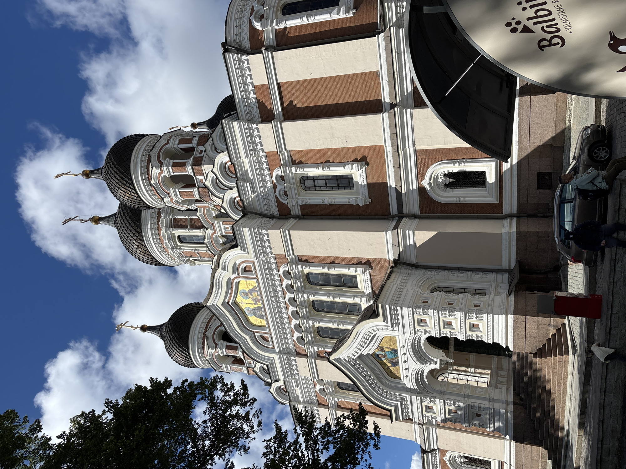The image shows a low-angle view of the Alexander Nevsky Cathedral in Tallinn, Estonia, on a partly cloudy day. The cathedral's distinctive red brick and cream-colored facade is visible, with its multiple onion domes topped with golden crosses. The foreground features a tree on the left, a set of stone stairs leading up to the cathedral's entrance, and a street with parked cars and a few people. A large, curved, dark awning extends from the right side of the frame, partially obscuring a circular sign with the word "Balbii" and a penguin graphic. One person is leaning against a low wall in the foreground, while another is sitting on the wall near a parked car. The sky is a vibrant blue with fluffy white clouds.