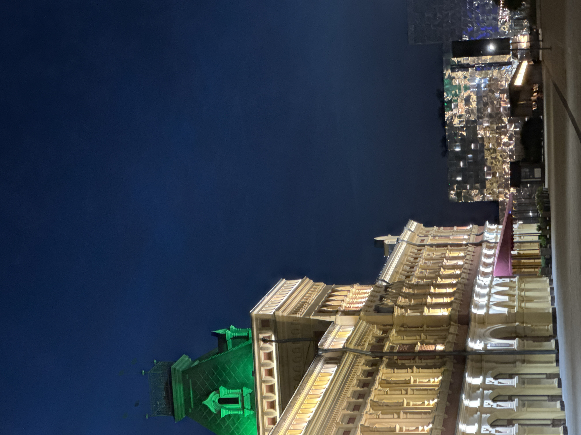 The image captures an exterior view of a building at night, with a dark blue sky as the backdrop. The building, illuminated by artificial lights, features a grand facade with ornate architectural details, including multiple levels of balconies and arched windows. The lighting highlights the building's texture and intricate design. To the left, a portion of another structure is visible, topped with a green, pitched roof and illuminated by green lights. Further in the distance, a modern building with a reflective, mosaic-like exterior is lit up, creating a contrasting visual element to the more traditional architecture. The scene suggests an urban setting at night, likely a city center or a well-lit commercial area. There are no people visible in the frame. The time of day is clearly night, and the sky indicates clear weather. The architectural styles suggest a historical European city, and the provided location context of Nizhny Novgorod, Russia, is consistent with such a scene.