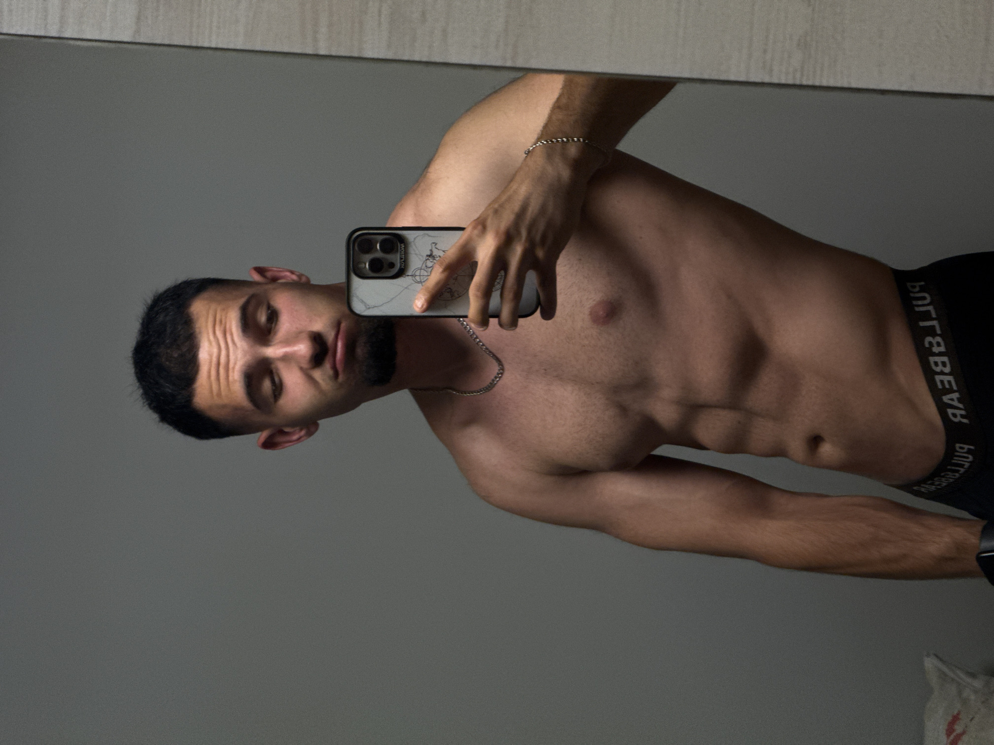A young man with short dark hair and a beard is taking a mirror selfie. He is shirtless, wearing black underwear with the brand name "PULL&BEAR" visible. He has a muscular physique with defined abs. He is holding a smartphone in his right hand, which has a marble-patterned case. A silver chain necklace is around his neck, and a silver bracelet is on his left wrist. The background is a plain gray wall and a portion of a mirror. The setting does not provide any specific visual cues to identify the city of Shumen, Bulgaria.