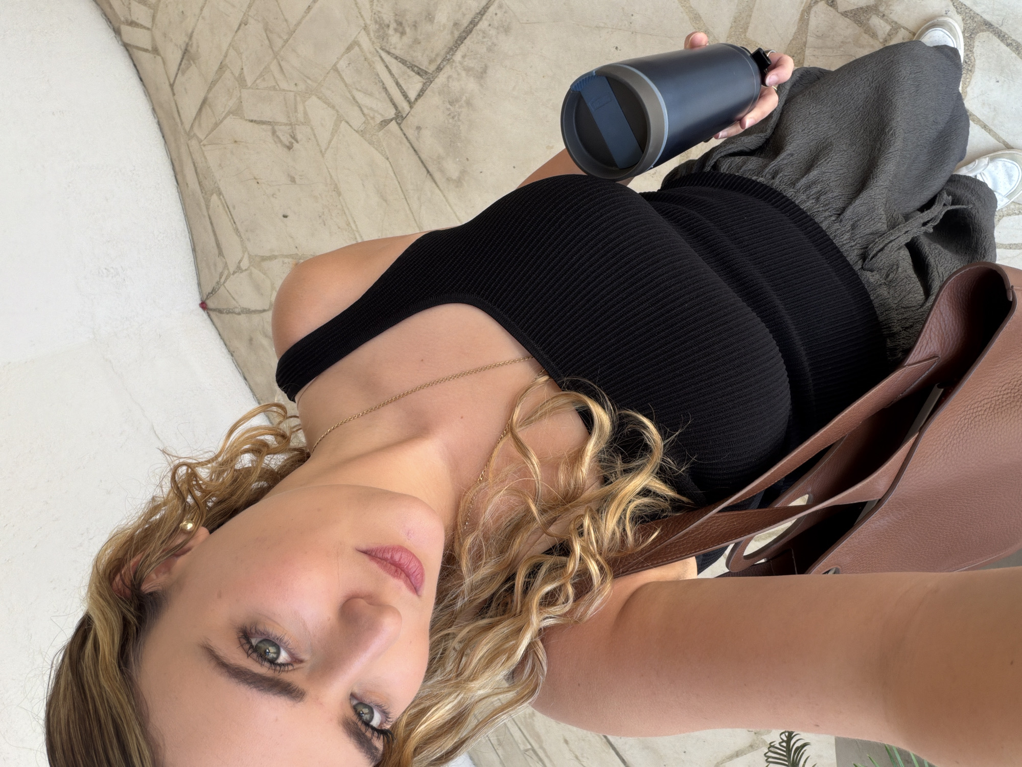 A high-angle, tilted photograph shows a woman in a casual outdoor or semi-outdoor setting, likely in San Bernardino Tlaxcalancingo, Mexico. She looks directly at the camera with a neutral to slightly smiling expression, highlighting her green eyes. Her wavy blonde and light brown hair cascades over her shoulders.
She is dressed in a black ribbed tank top or dress, paired with dark grey or olive green textured, loose-fitting pants, and white sneakers. Accessories include a thin gold chain necklace and a gold stud earring. In her right hand, she holds a dark blue or black reusable water bottle with a grey lid. A brown leather shoulder bag is visible on her left side.
The background features light-colored, irregularly shaped tiled flooring, possibly flagstones, and a plain white, textured wall or surface. The bright, natural light suggests the photo was taken during the daytime. The woman appears to be posing for the shot. No legible text is visible in the image.