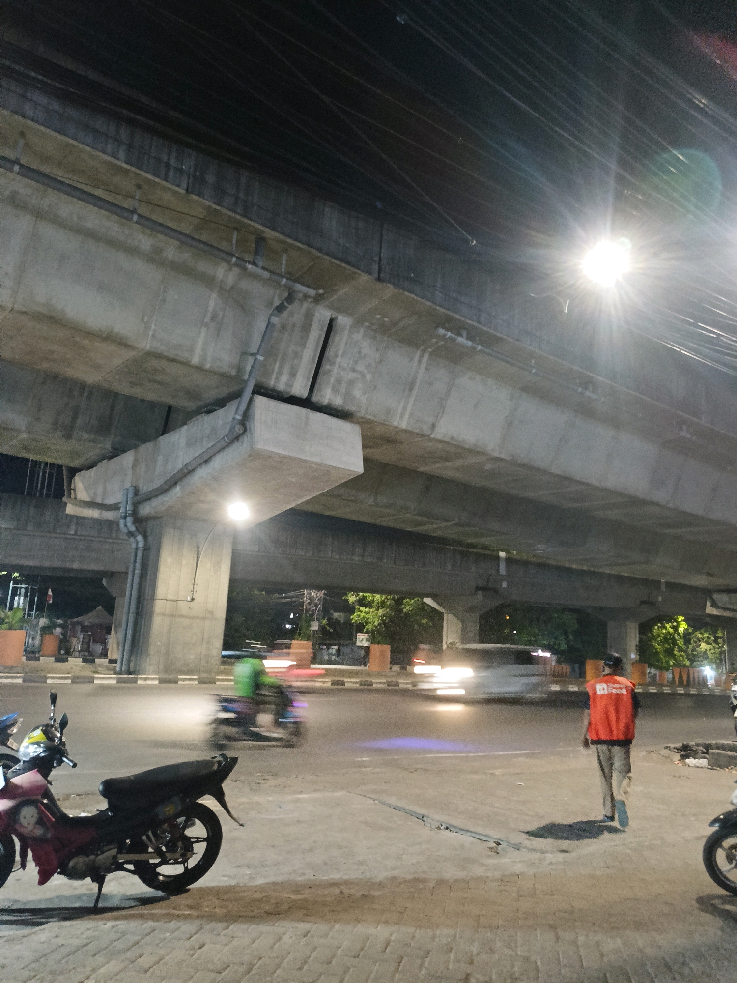The image depicts a street scene at night in Makassar, Indonesia, beneath a large concrete overpass. The overpass, a prominent structure, casts shadows on the road below. The scene is illuminated by streetlights, one of which is a particularly bright, starburst-like light source in the upper right quadrant of the image.
In the foreground, a red and black motorcycle is parked on the side of the road, facing left. Its details are relatively clear, with visible wheels, handlebars, and a seat.
On the road, vehicles are in motion, indicated by motion blur. A person wearing an orange vest and light-colored pants walks away from the camera towards the right side of the frame. The person is the most clearly depicted human subject in the scene. Further down the road, a green-clad figure on a motorcycle is also visible, blurred due to movement.
The environment suggests an urban setting, with trees and some temporary structures or stalls visible in the background. The road surface appears to be paved with interlocking bricks in the foreground. The overall impression is that of a bustling urban intersection at night, possibly with ongoing construction or infrastructure development due to the presence of the large overpass.