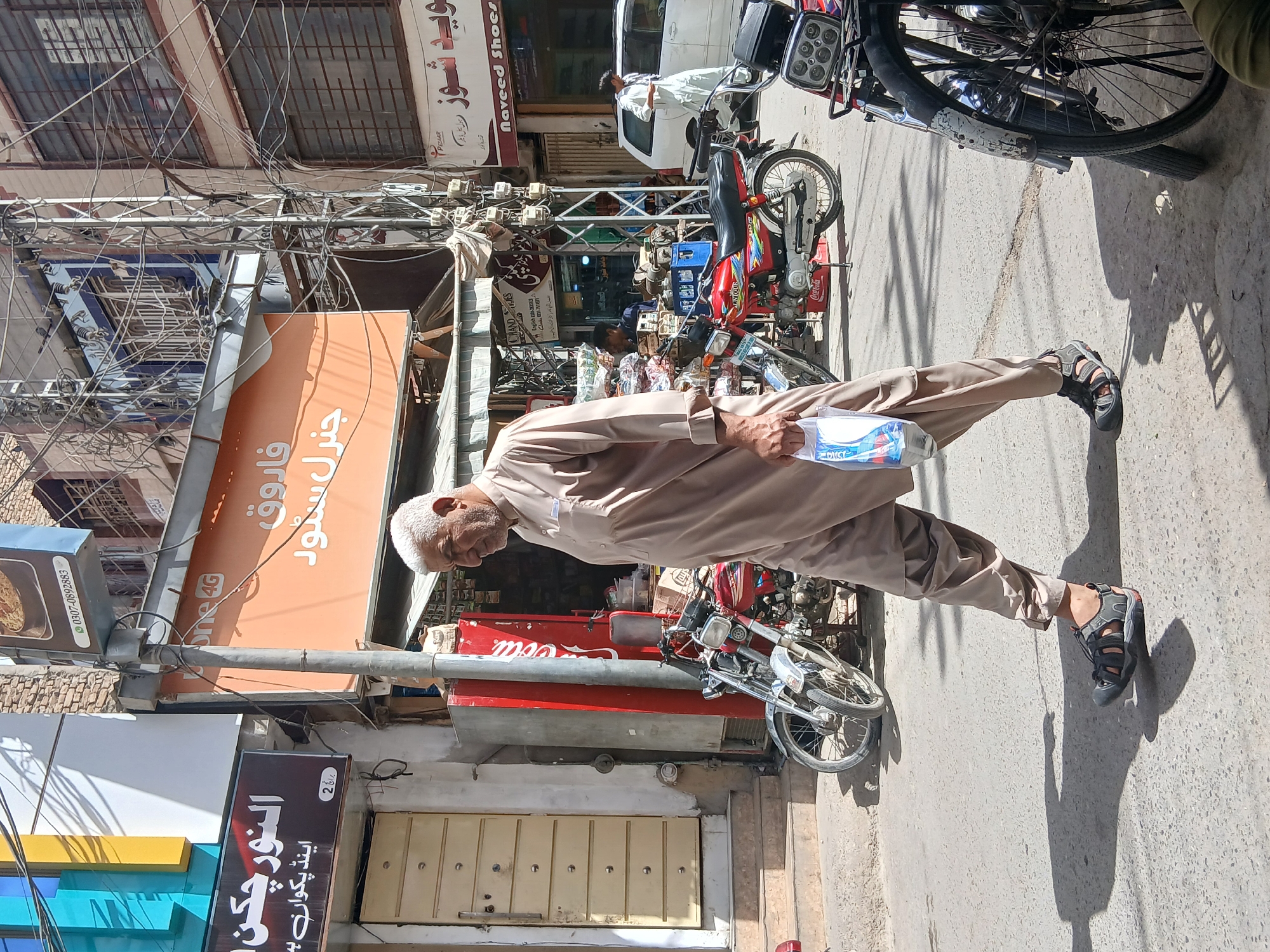 An elderly man walks along a street in Quetta City Tehsil, Pakistan. He is wearing a light brown traditional outfit and sandals, carrying a package in his left hand. Behind him are several parked motorcycles and commercial buildings with signs in Urdu. Overhead, a tangle of wires and cables adds to the busy street scene.