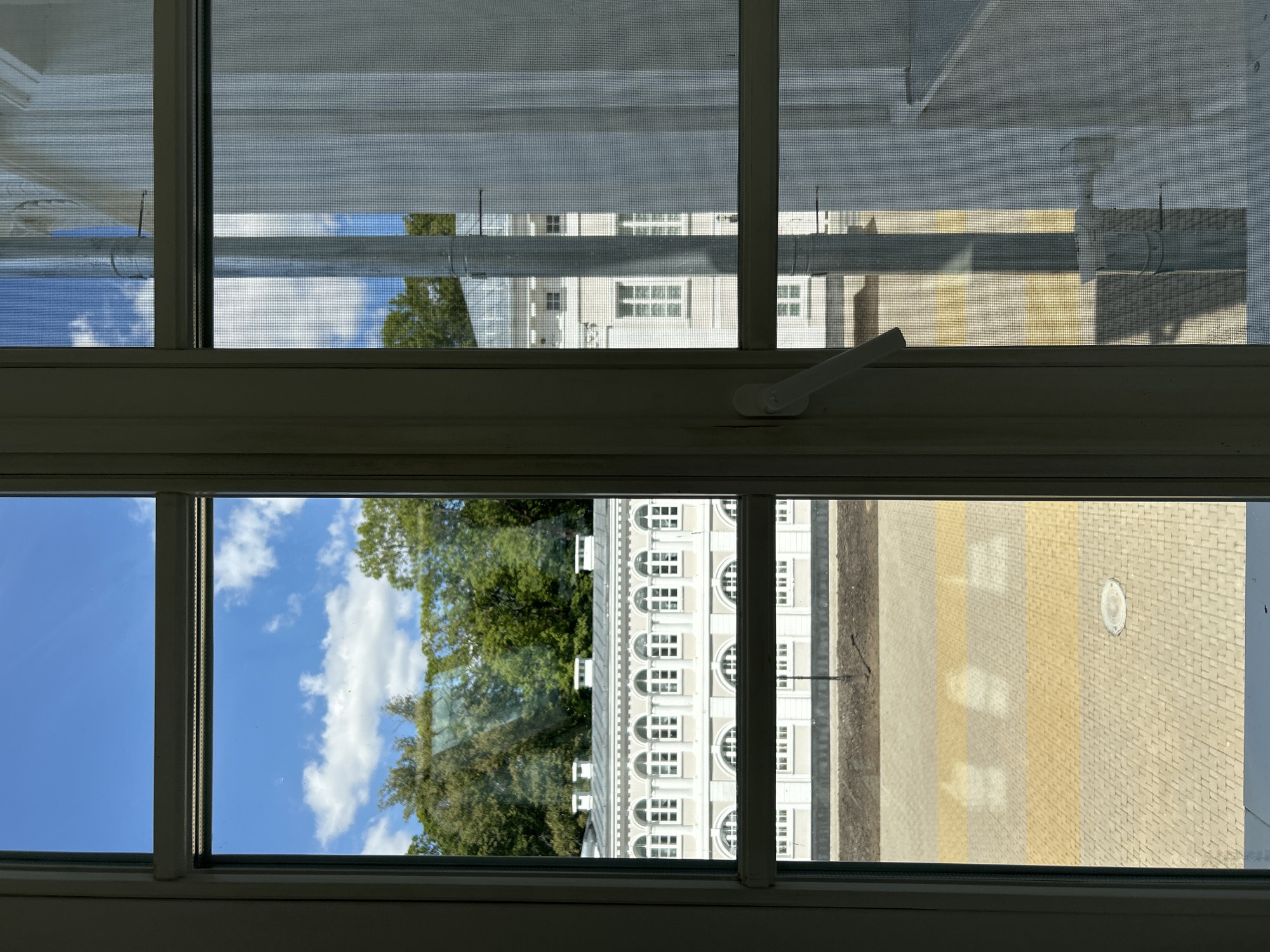 The image shows a view through a window, looking out at a white building with arched windows and a tiled roof, surrounded by trees. A paved courtyard with yellow and grey rectangular pavers is visible in the foreground. The sky is blue with scattered white clouds. The scene is set in the city of Dobasnenskі sel'ski Save, Belarus.