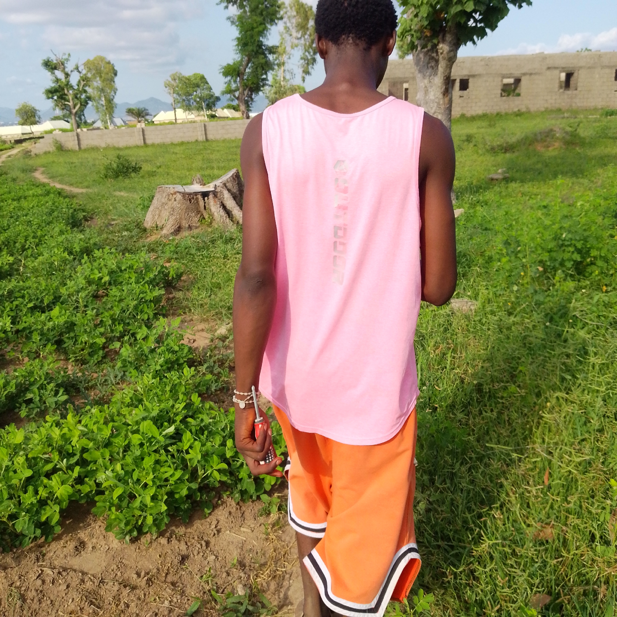 A person is walking away from the camera in an outdoor setting with lush green vegetation and a building in the background. The person is wearing a pink tank top and orange shorts with white and black trim. The person is also wearing bracelets on their left wrist and is holding a screwdriver. The ground is a mix of dirt and grass, and there are trees in the background. The building appears to be under construction and has a concrete facade. The sky is partly cloudy.