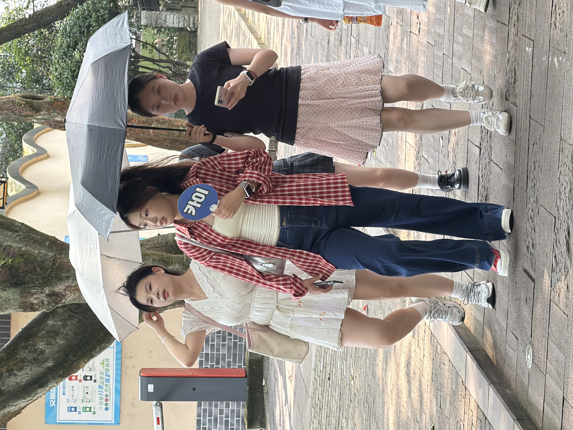 The image depicts four young women walking on a paved pathway outdoors in Yuzhong District, China.
From left to right, the first woman, in her early 20s, wears a white short-sleeved dress with lace trim at the V-neck and a thin, decorative belt. She carries a light pink shoulder bag and has short dark hair, with her right hand positioned near her head. She wears silver athletic shoes with white socks. Adjacent to her, an automatic barrier arm with dark gray and red sections is visible on the far left. Above and behind her, a blue rectangular sign displaying waste classification graphics and Chinese text related to environmental protection is mounted on a wall.
The second woman, also appearing to be in her early 20s, wears a red and white plaid long-sleeved shirt unbuttoned over a light-colored tank top, paired with dark blue wide-leg jeans. She holds a blue circular fan with the white Korean characters "카이" (KAI) in front of her chest. Her dark hair is pulled back, and she wears white shoes with red toe accents. A silver shoulder bag is visible behind her right arm.
The third woman, in her late teens or early 20s, holds a black umbrella above her head with her left hand. She is dressed in a black short-sleeved t-shirt and a light pink skirt with small dark polka dots and a pleated hem. She looks down at a light-colored smartphone held in her right hand and wears white athletic shoes with white socks.
A fourth woman is partially visible behind the second woman, wearing a dark top, a dark plaid skirt, and black chunky loafers.
The pathway is constructed of rectangular paving stones. In the background, large trees with green foliage and a light-colored building featuring a scalloped roofline are present. The scene is illuminated by natural daylight.