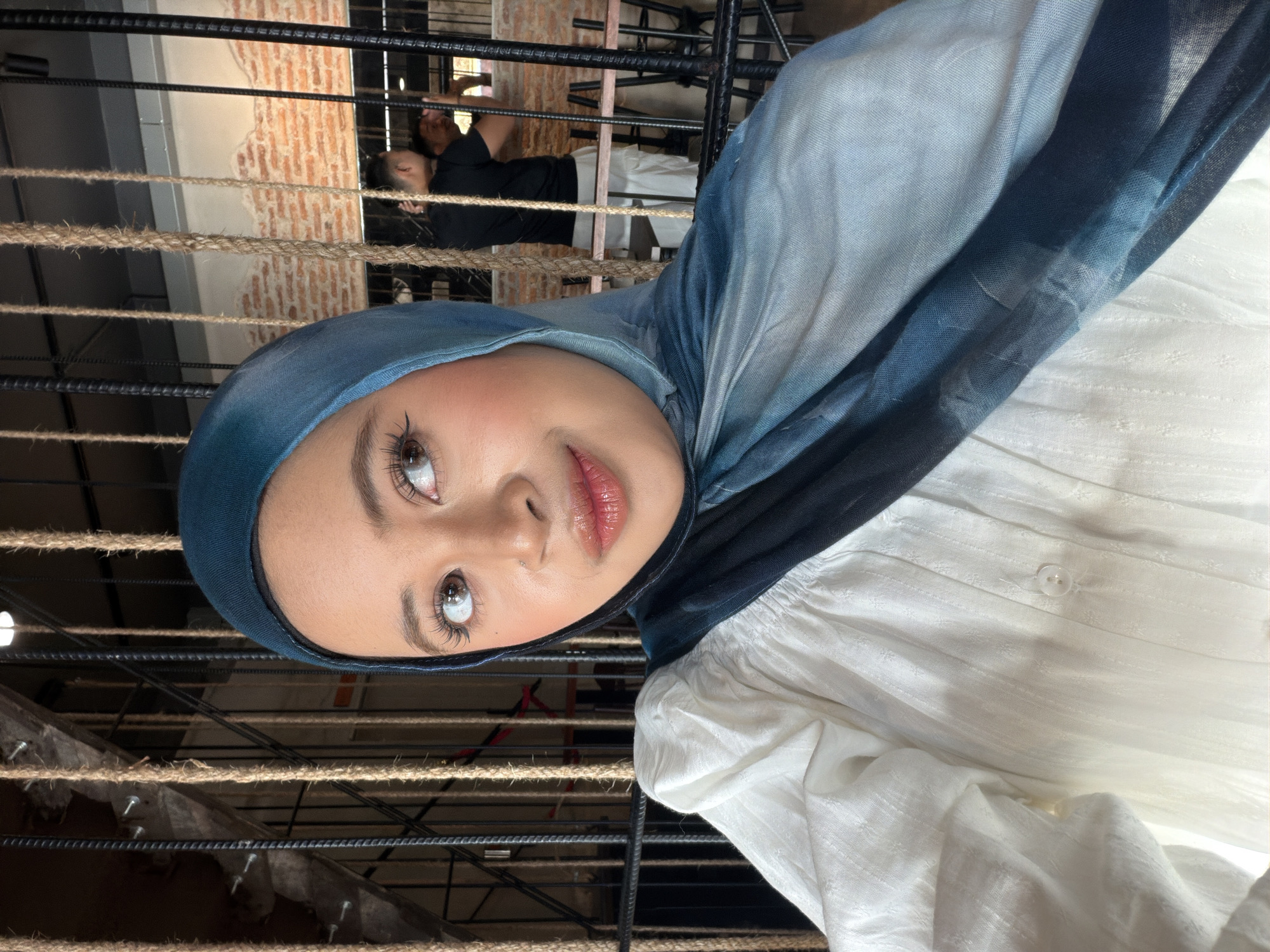 An individual wearing a hijab and white blouse poses for a selfie in an indoor setting. The background consists of rope and metal structures, with exposed brick walls and other patrons visible in the distance. The lighting suggests an ambient indoor environment, possibly a cafe or restaurant. The subject's makeup is noticeable, with emphasis on the eyes and lips. The image appears to be taken in Bangkok, Thailand, based on visual cues.