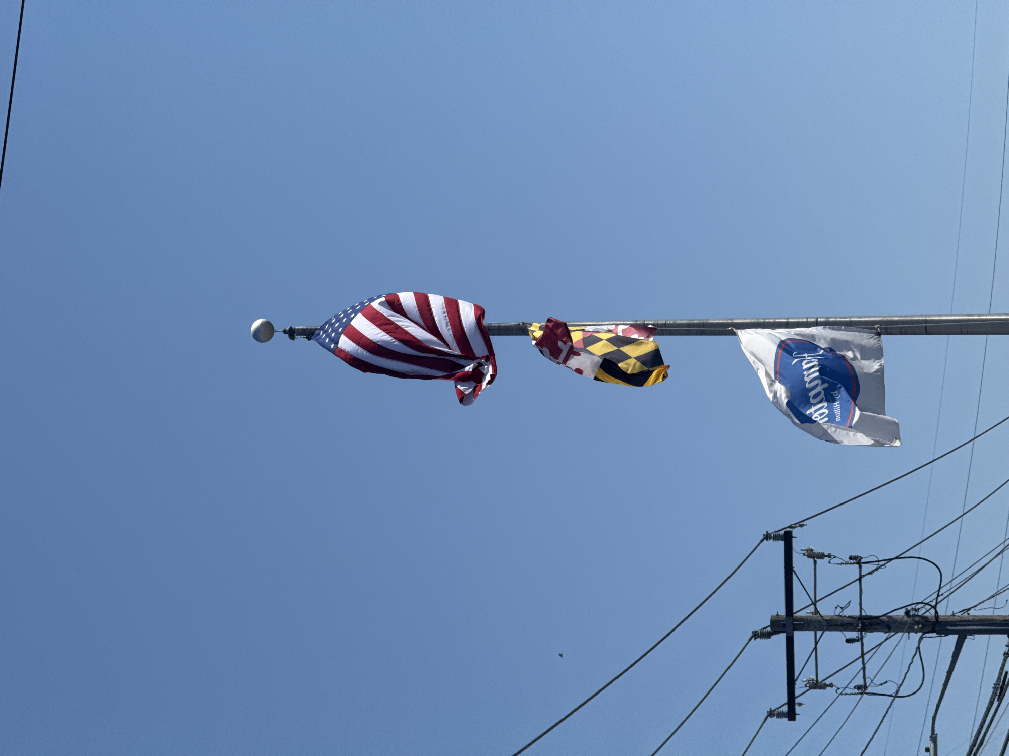 The image shows a low-angle view of three flags attached to a horizontal pole against a clear, blue sky. From left to right, the flags appear to be: the flag of the United States of America, the flag of the state of Maryland, and a white flag with a blue emblem and text, that is hard to make out. Utility poles with numerous power lines are visible in the lower right quadrant. Given the presence of the Maryland state flag and the utility infrastructure, the location is likely to be a neighborhood or commercial area somewhere in the city of Ocean City, United States of America.