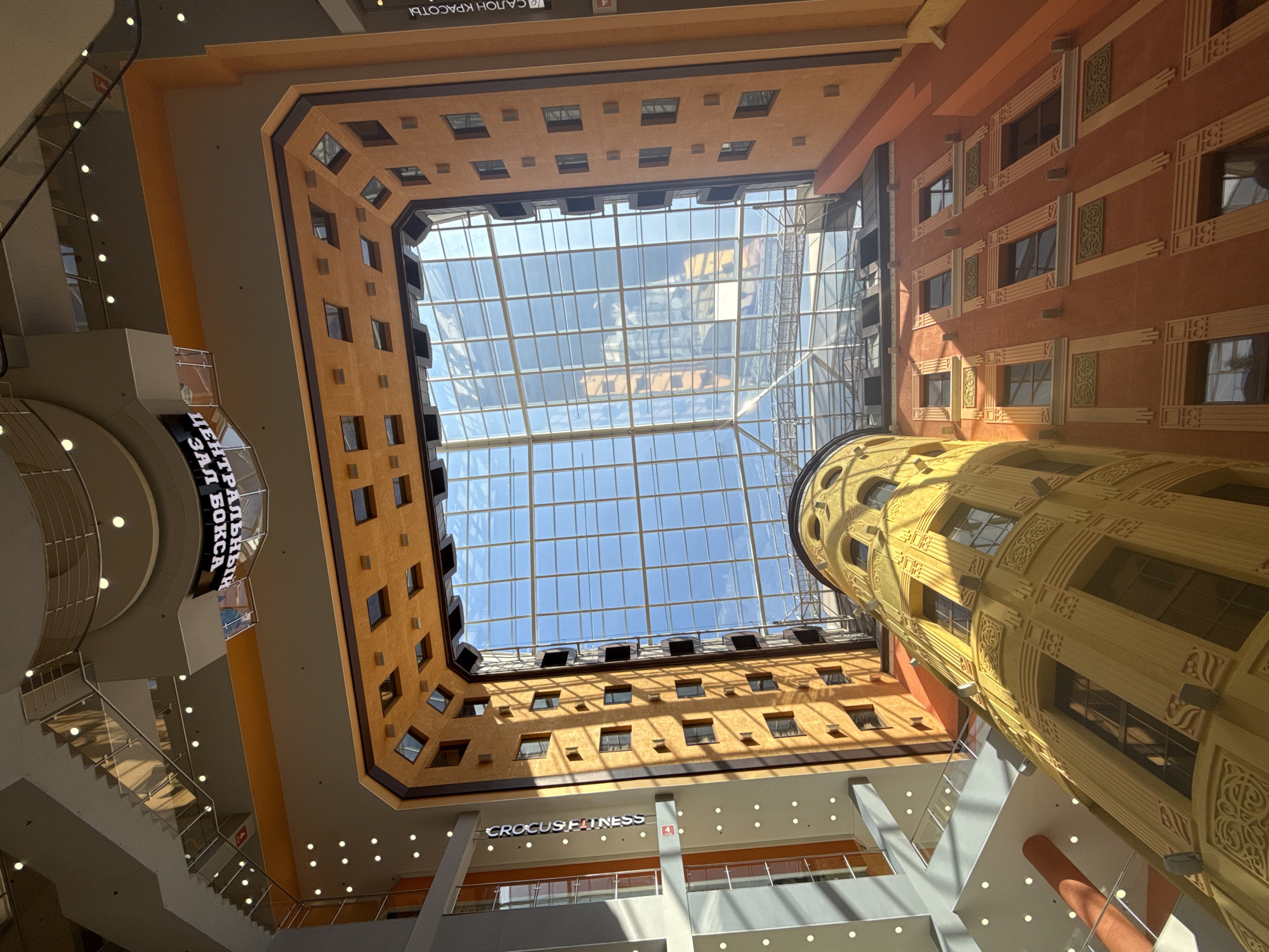 The photograph captures the bright, multi-story interior of what appears to be a large commercial or recreational complex, likely located in Saint Petersburg, Russia. The perspective is looking upwards into a vast central atrium.
The most prominent feature is a large, rectangular glass skylight that forms the ceiling, allowing abundant natural sunlight to illuminate the space. The sky, visible through the glass, appears clear and blue. Shadows cast across the lower levels indicate direct sunlight, suggesting it is daytime.
The inner facades of the building are rendered in warm earthy tones, primarily orange and light brown/tan, with numerous small, dark-paned windows arranged in regular patterns. One side features more elaborate decorative elements around the windows and along the walls. A striking, tall, cylindrical tower, painted in a vibrant yellow or gold and adorned with intricate, art deco-inspired patterns and numerous windows, stands prominently on the right side of the atrium. Multiple levels are visible, connected by escalators on the left side of the frame, which are illuminated by recessed lights.
No people are visible in the image. The setting suggests a bustling public space, possibly a shopping mall, exhibition center, or a multi-purpose complex.
Visible signs and text further clarify the building's purpose:
- "CROCUS FITNESS" is clearly displayed in white letters on a dark background on one of the lower levels, indicating a fitness center.
- Partially visible Cyrillic text includes "САЛОН КРАСОТЫ" (SALON KRASOTY - Beauty Salon) on a higher floor.
- Another sign, partially obscured, reads "ЦЕНТРАЛЬНЫЙ ЗАЛ БОКСА" (TSENTRALNYY ZAL BOKSA - Central Boxing Hall).
- A red square with the number "4" is visible, likely indicating a floor level.
The overall impression is one of a spacious, well-lit, and architecturally interesting modern interior, designed to accommodate various commercial and recreational activities.