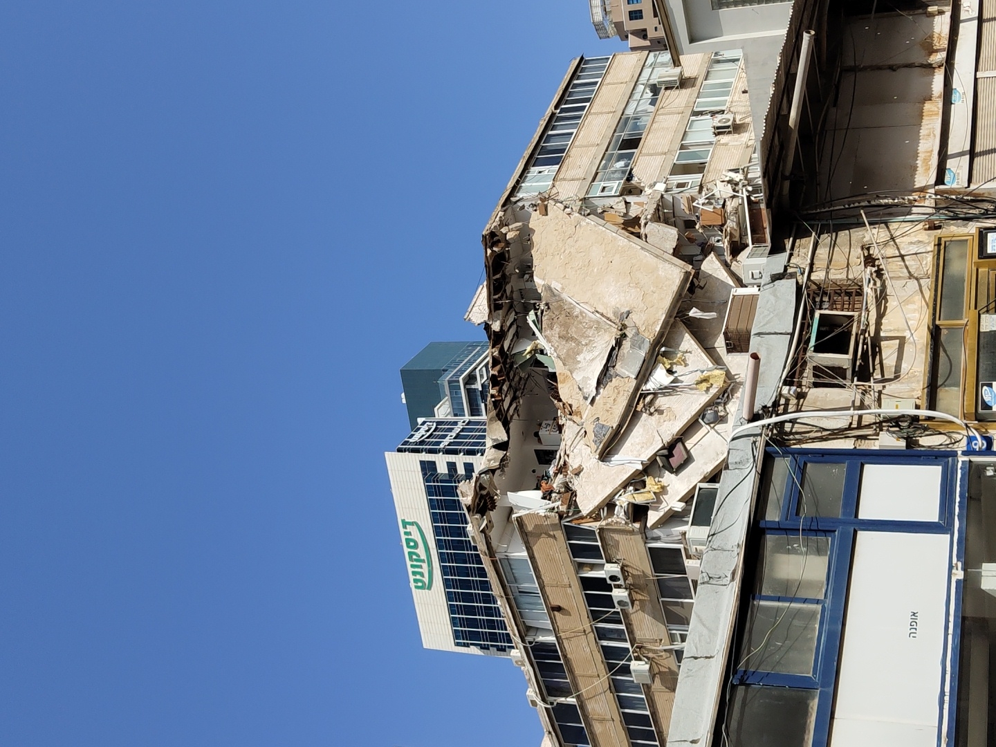 The image shows a collapsed building in Tel-Aviv, Israel. The building's facade is heavily damaged with exposed concrete and debris. A neighboring building with a sign in Hebrew, "דירה," is visible. Debris is scattered around the base of the destroyed building, and a clear blue sky is visible in the background. Electrical wires and other infrastructure are visible in the foreground. No people are visible in the frame.