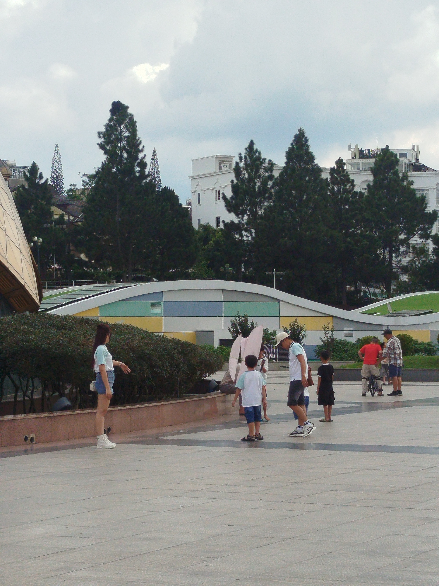 The image depicts an outdoor scene in a public space in Da Lat, Vietnam, likely near a modern architectural structure with a curved roof and colorful panels. Several individuals are present, including adults and children.
In the foreground, a young woman in a light blue and white t-shirt and denim shorts stands with her back to the camera, facing to the right. She is wearing white boots. To her left, a small group of children is visible. One child, wearing a light blue t-shirt and blue shorts, is standing and looking towards the right. Another child, in a black t-shirt and shorts, is also present.
Further back, a man in a white t-shirt, dark shorts, and a white cap is interacting with a child. He is carrying a brown bag. To his right, a man in a red t-shirt and beige shorts is standing near a child on a unicycle. Another man in a checkered shirt and dark shorts is standing to the right of the red-shirted man, holding a smaller child's hand.
The background features lush green trees, and buildings. The sky is overcast with clouds. The ground is paved with large, grey square tiles. A low, brown wall borders a raised garden area with dense green bushes. The overall atmosphere suggests a casual gathering or outing in a city park or plaza.