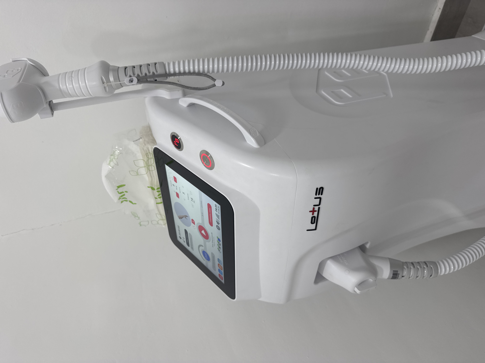 The image shows a Lotus-branded medical device, possibly a laser hair removal machine, mounted on a white wall. The device is primarily white with a digital display, a handle, and several attached hoses and wands. The setting appears to be indoors. There is no indication of the location being in Kuta Selatan, Indonesia, or any indication of people, actions, or interactions in the scene.