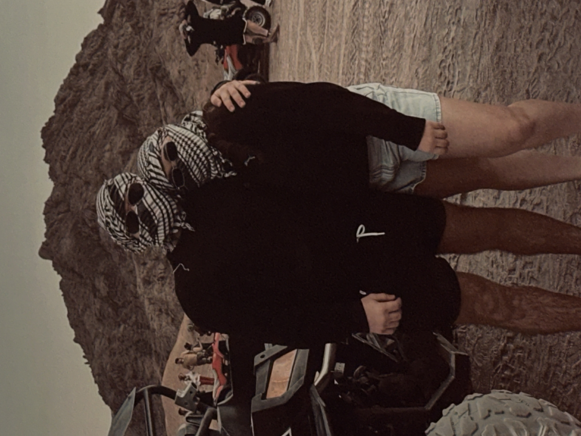 Two people, partially obscured by keffiyehs and sunglasses, embrace in a desert environment. The person on the left wears a black sweatshirt and black shorts, while the person on the right wears a black sweatshirt and light denim shorts. They stand in front of a mountainous, rocky landscape. In the background, several ATVs and other people are visible, suggesting an outdoor recreational activity. The ground is sandy and uneven, marked with tire tracks. The sky is overcast and hazy.
