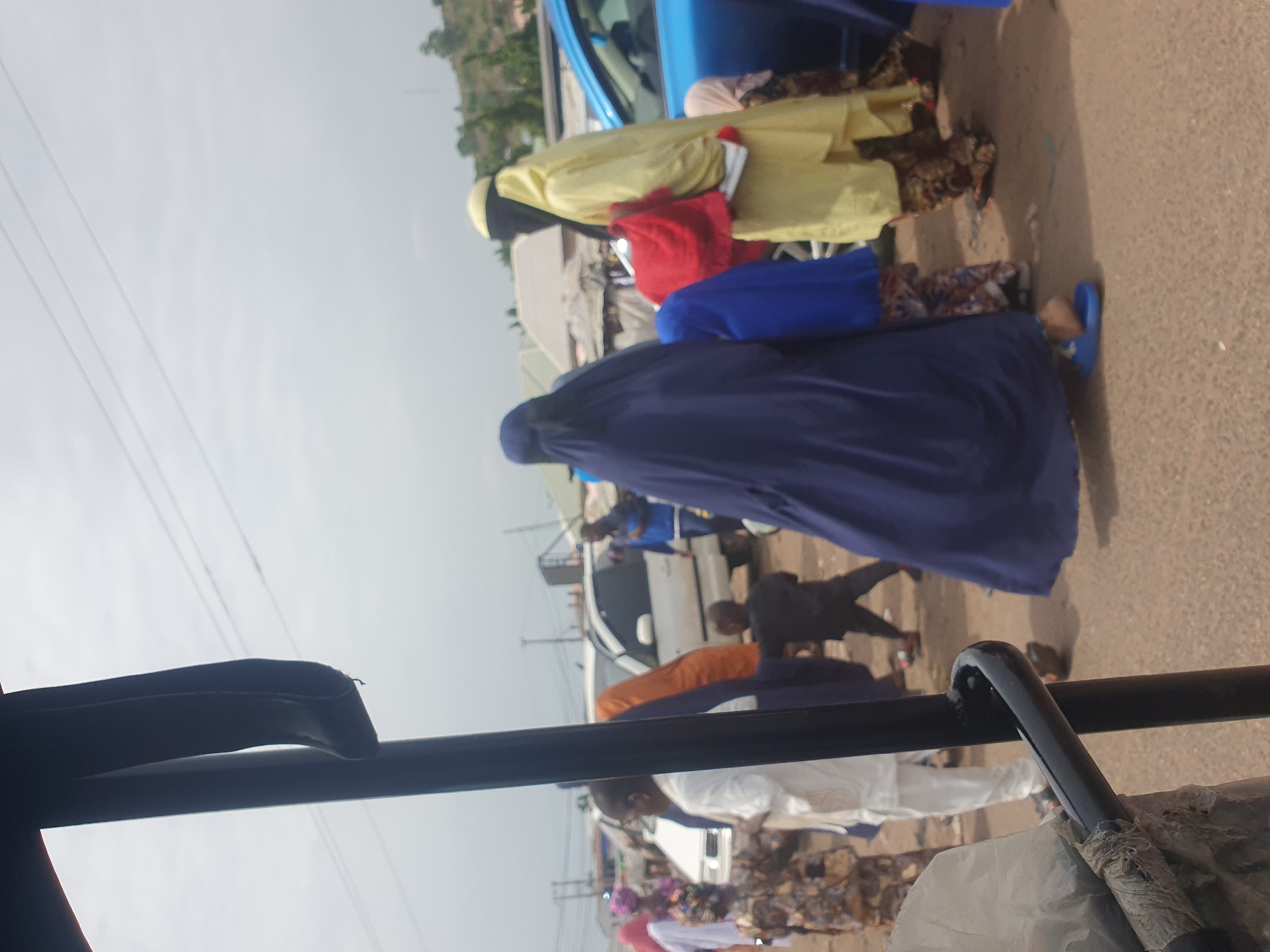A ground-level, medium-shot shows a street scene in Lokoja, Nigeria. The image is shot from inside a vehicle, through its black metal frame.
Several individuals are visible on the street. A group of people, mainly women, are dressed in brightly colored traditional Muslim attire including a navy blue, yellow, and red garment. They are standing near a blue car parked on the side of the street. In the background, other individuals walk or stand among parked cars and roadside structures. The environment appears to be a bustling urban area. Overhead power lines are also visible against a pale sky.