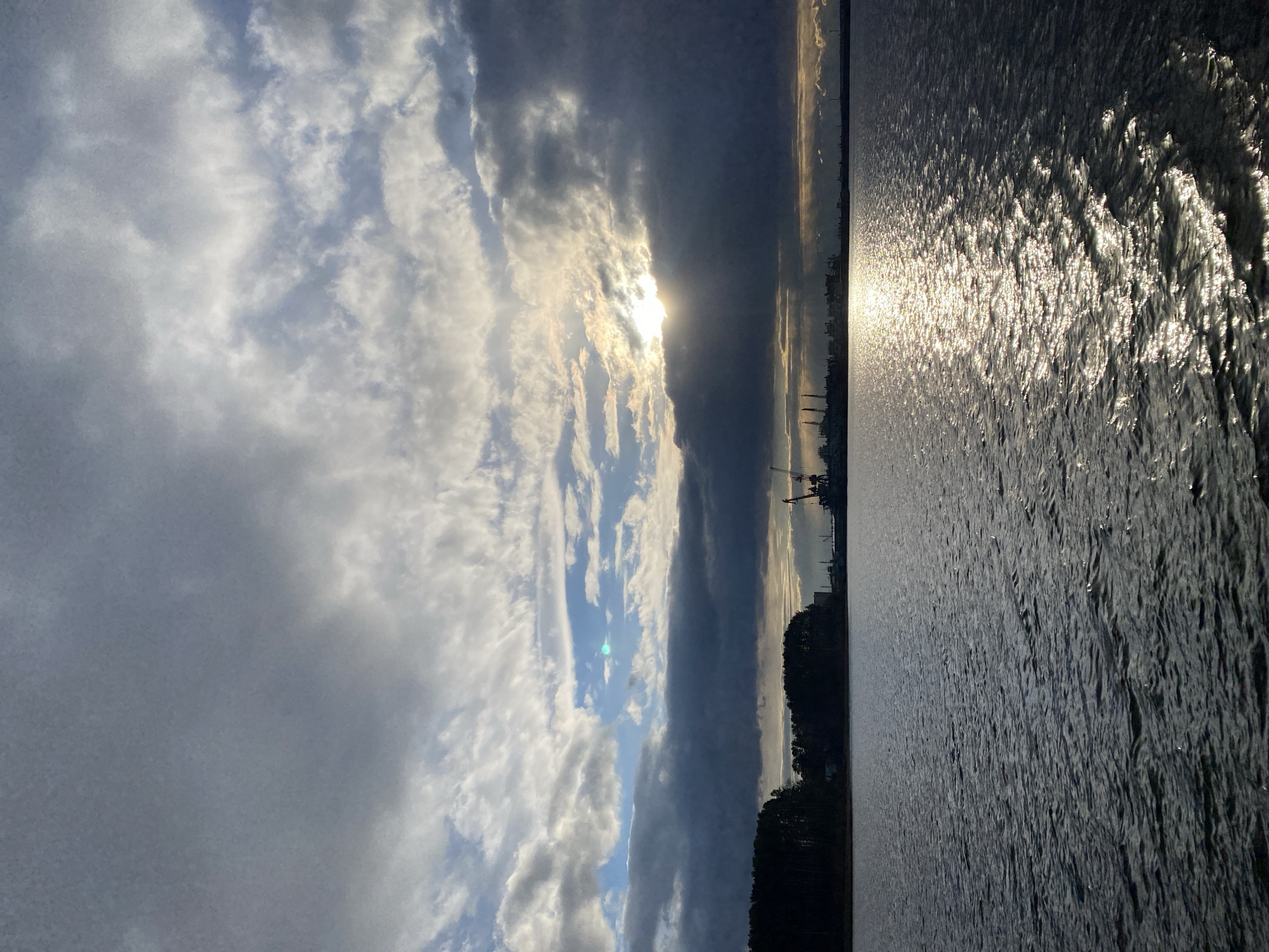 The image, when correctly oriented, depicts a dramatic waterscape scene, likely at sunrise or sunset, in Chaikovsky, Russia. The sky fills the upper portion, showcasing a dynamic mix of voluminous dark and brightly lit clouds, with the sun partially obscured but emitting a powerful light. This light creates an intense, shimmering glare and textured patterns across the surface of the wide body of water that dominates the lower half of the frame. Along the distant horizon, a dark, undulating landmass, possibly forested, is silhouetted. Above this land, faint outlines of industrial structures, resembling cranes or port facilities, are visible against the lighter sky, indicating human presence. The strong contrasts between bright highlights and deep shadows contribute to a powerful and expansive atmosphere. No text is visible in the image.