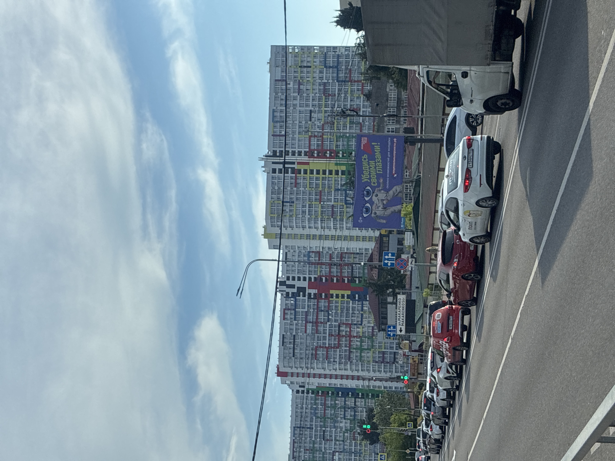 The image shows a traffic jam on a sunny day in Sochi, Russia, with large, colorful apartment buildings in the background. The buildings are modern, with many windows and balconies, and are painted in a variety of bright colors, including white, blue, red, green, and yellow. There is a large billboard with an advertisement for what appears to be an optical store. The road is filled with cars, and the traffic appears to be moving slowly. There are traffic lights and street signs visible, including one that reads "ул. Макаренко MAKArenko Str.". The sky is blue with some white clouds. The overall scene depicts a typical urban environment with a significant amount of vehicle traffic.