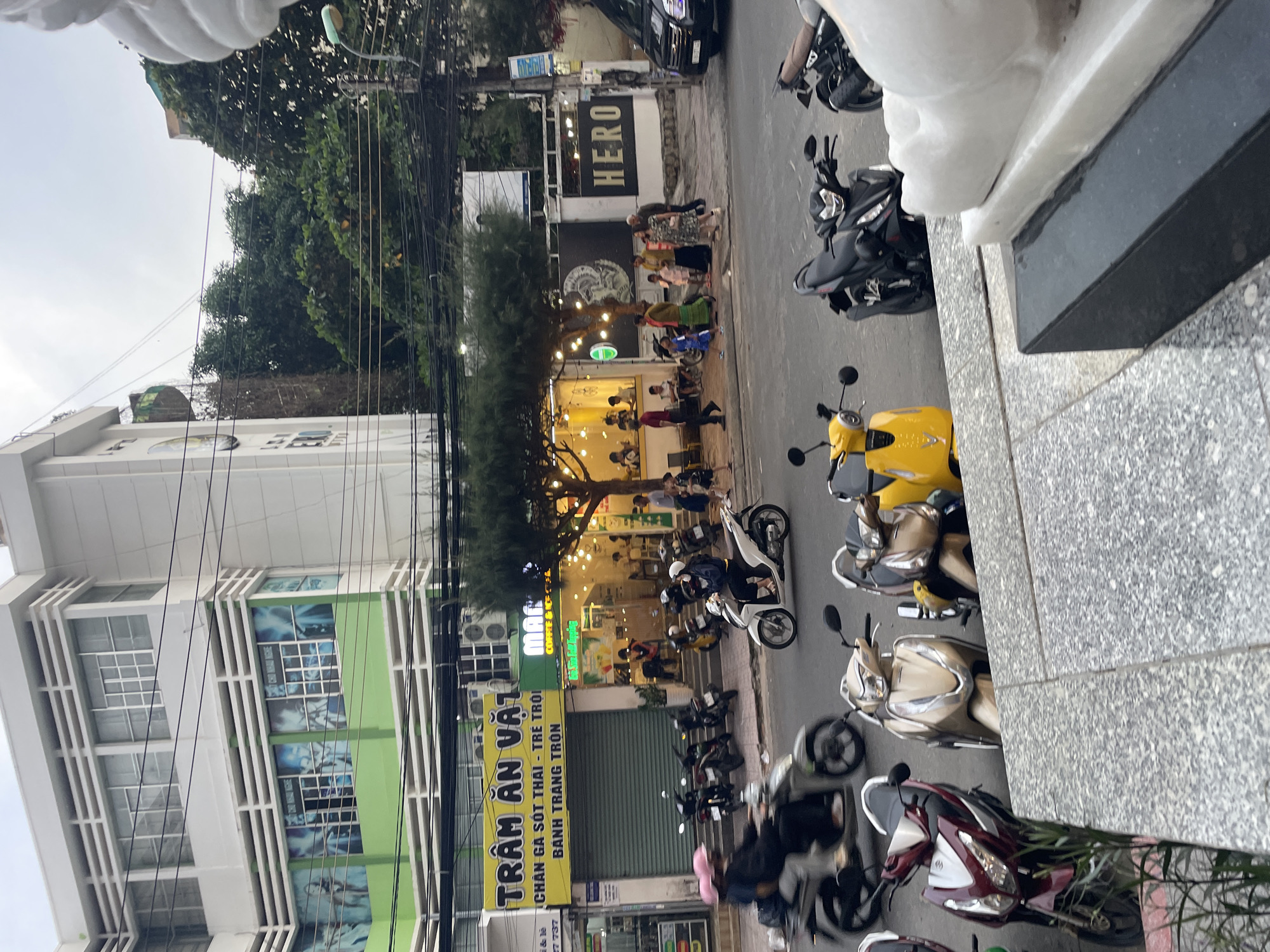 The image depicts a street scene in Bac Nha Trang Ward, Vietnam. In the foreground, several parked motorbikes are visible. One is a dark red scooter, a gold scooter, and a yellow scooter. Further back, a white scooter is being ridden by a person wearing a dark blue jacket and a white helmet. Another scooter, a dark scooter, is also visible with a person wearing a pink helmet.
To the left, a building with a green lower section and white upper section features large windows with blue graphics. Text on a yellow sign above a storefront reads "TRÂM ĂN VẶT" followed by smaller text that appears to be a menu. Adjacent to this is a store with green signage and the word "MAGIC" visible, with "COFFEE & ICE TEA" below. This store is illuminated with string lights and has patrons seated inside.
Across the street, there is a white wall with a dark graphic of a classical bust and the word "HERO" in large, dark letters. A dark vehicle is parked to the right of this wall. The environment appears to be urban, with overhead power lines and trees lining the street. The sky is overcast.
