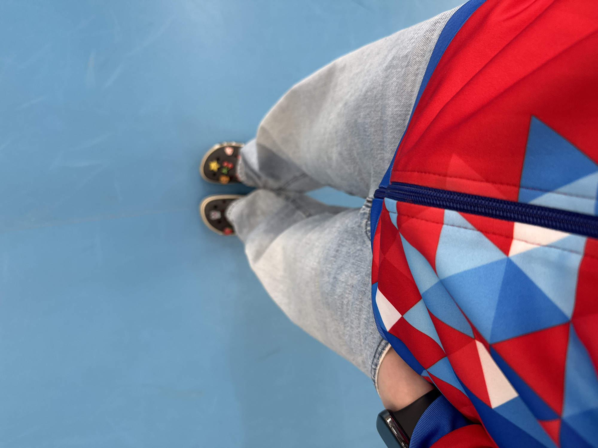 A top-down shot shows a person standing on a light blue surface, likely a floor. They are wearing light-wash jeans and black slip-on shoes with colorful embellishments on the toes. Part of a red, blue, and white geometrically patterned jacket is visible on the right side of the frame. A black smartwatch is visible on the left wrist. The scene is likely indoors given the even lighting. No specific information is available from the image alone to verify it is Usolye, Russia.