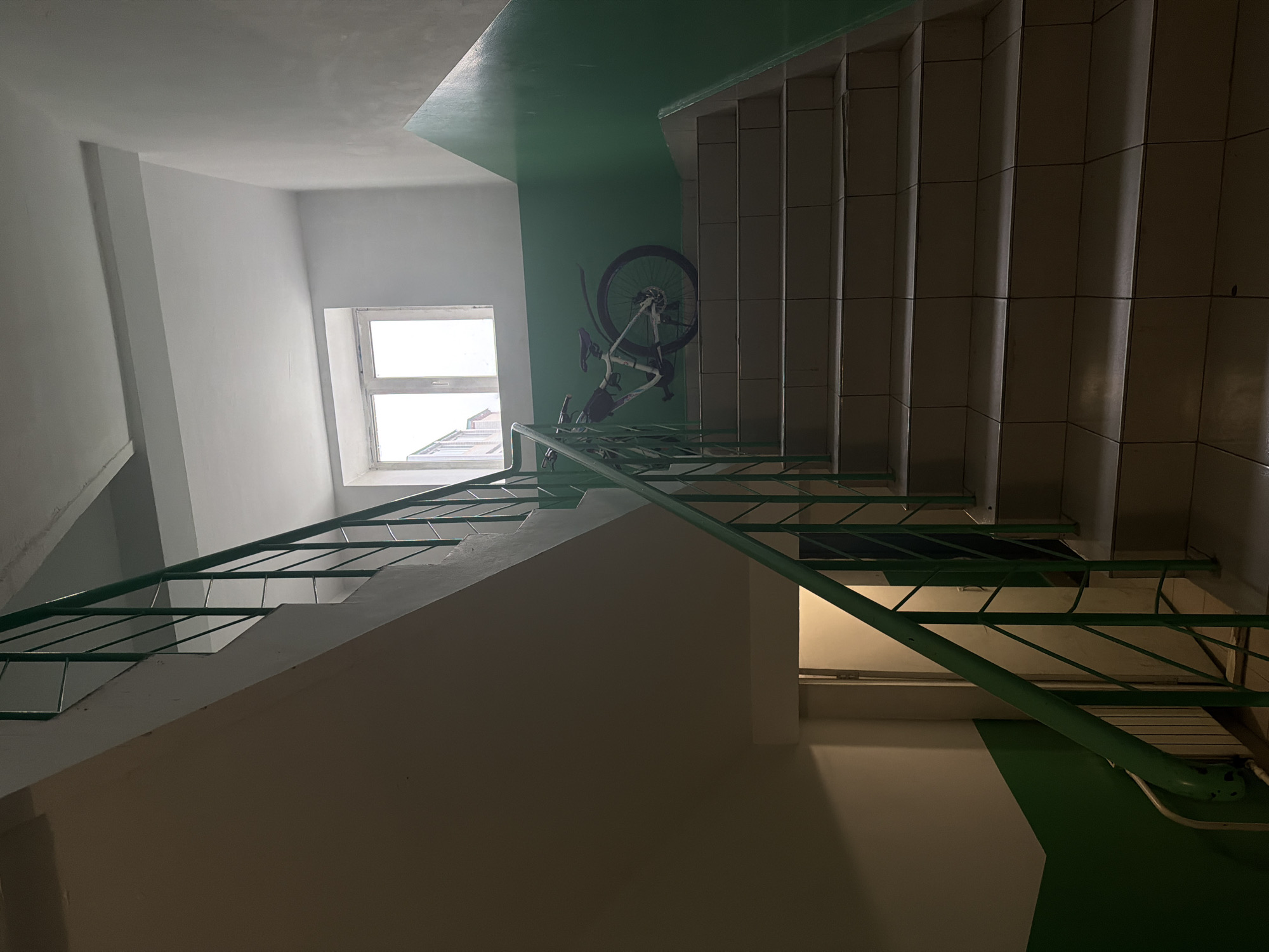 The image shows an interior view of a stairwell, likely in an apartment building. The walls are painted white, except for a section painted green on the upper level. A bicycle is hung on the green wall. A window provides natural light. The stairs are concrete with tile accents, and have green metal railings. The location is in an unidentified building in Birobidzhan, Russia. No people or subjects are visible.