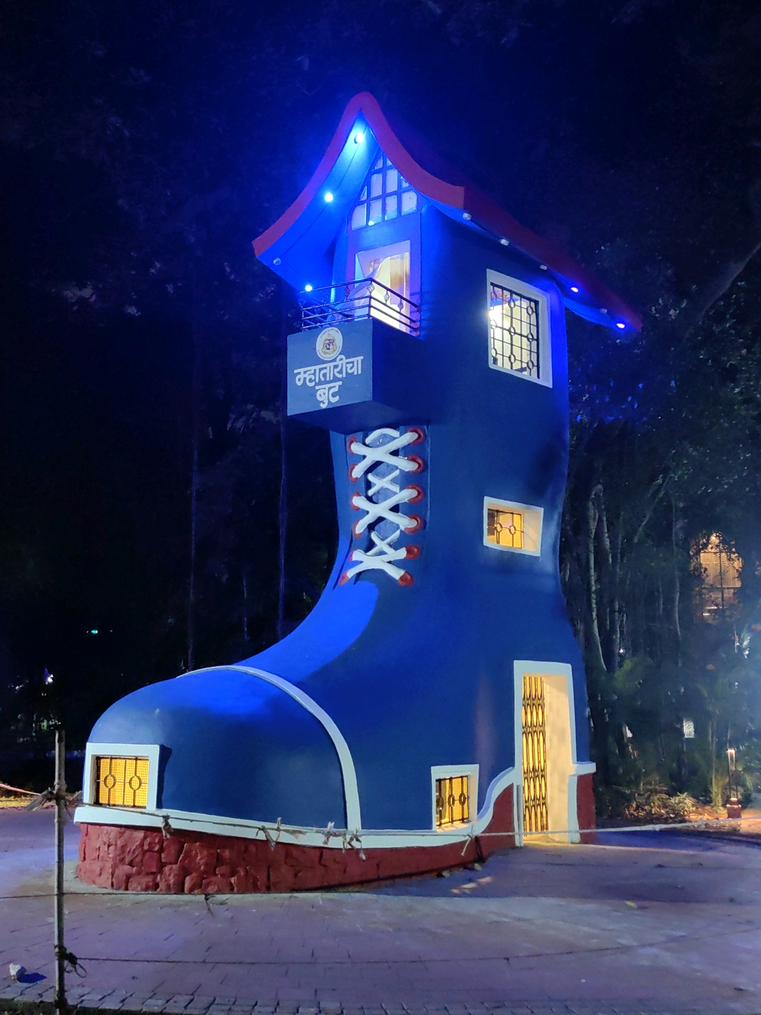 The image shows a large, blue boot-shaped building at night, illuminated by blue and white lights. The building has a red base and a red-and-blue roof. There are windows on the boot, some of which are lit with a warm yellow light. A door with a decorative metal gate is visible on the right side of the building. There is text in Marathi on a blue sign above the door, which appears to read "महातरीचा बुट" (Mahatricha Boot). The building is surrounded by trees and foliage, and the ground is paved with stone. A metal pole with wires is visible in the foreground on the left. This structure is located at Kidzee School, Kandivali West, Mumbai, India, and is part of a garden area.