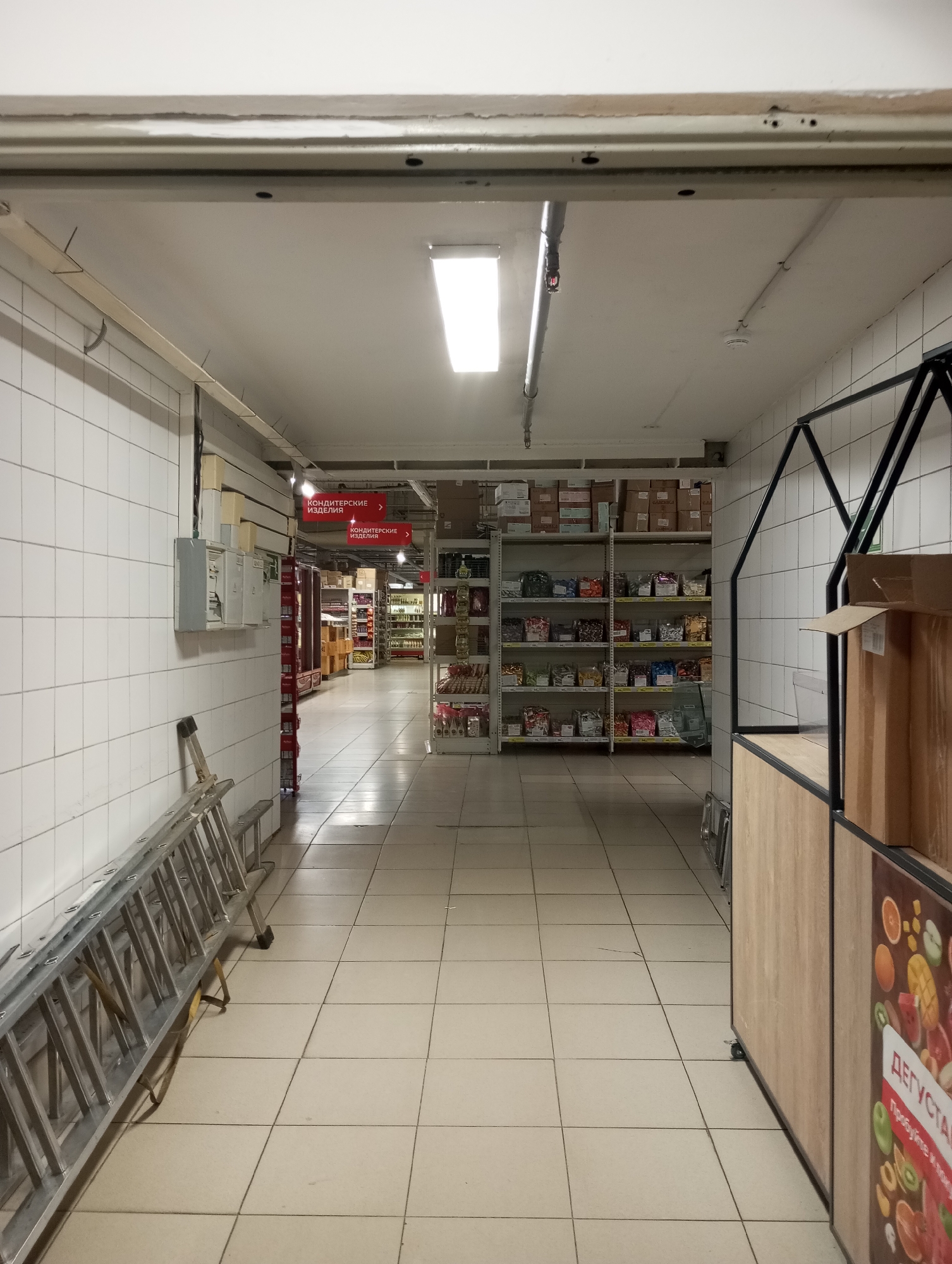 Aisle in a grocery store in Kamensk-Uralsky, Russia. The aisle has white tiled walls and floor. A metal ladder leans against the left wall. Shelves stocked with items are visible along the right wall. Red signs in Russian point towards "Confectionery". A display case stands on the right, partially obscured by boxes. Overhead, fluorescent lights illuminate the scene.