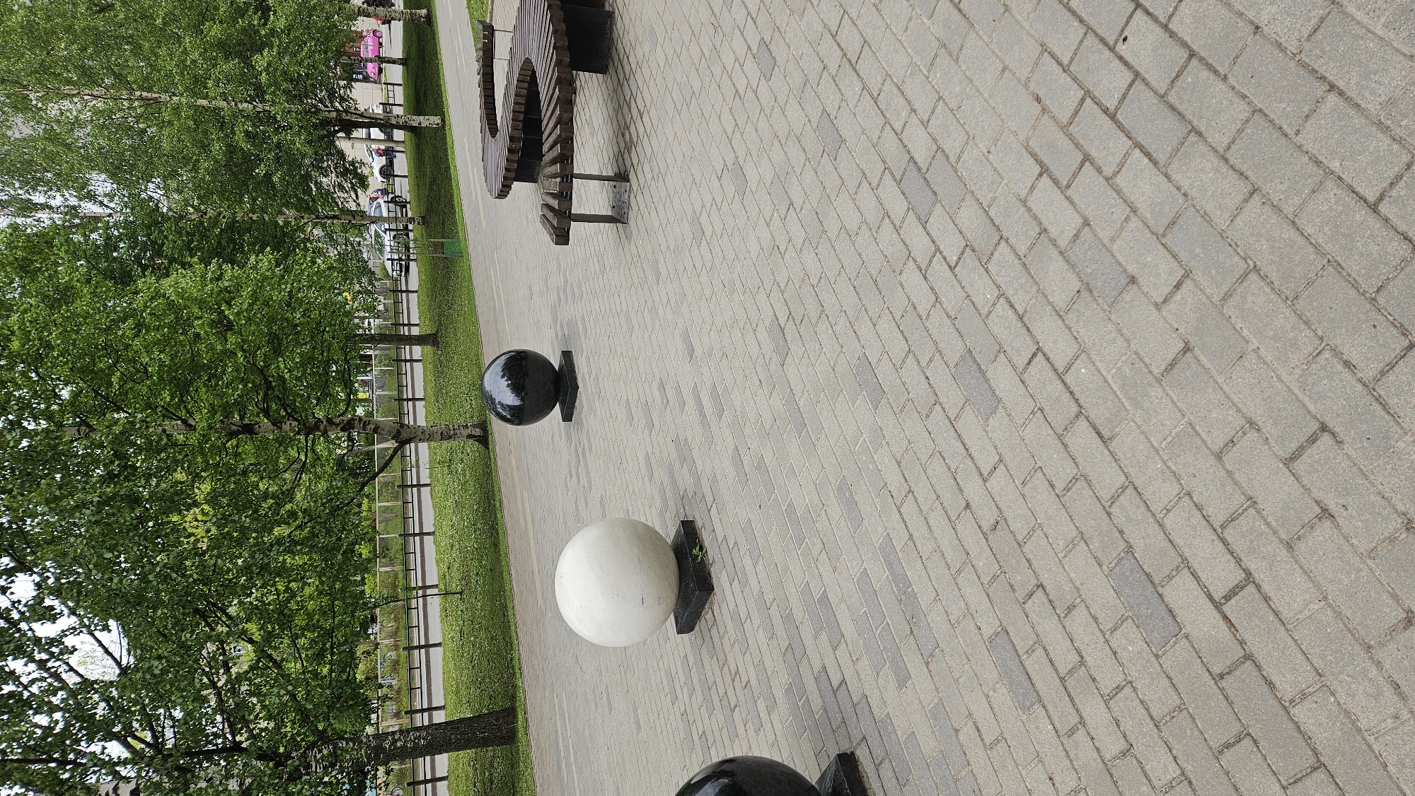 The image shows a paved walkway in an urban setting. The walkway is composed of gray rectangular pavers laid in a running bond pattern. Along the left side of the walkway are trees with green foliage and a grassy area. A black metal fence separates the grassy area from the background. Scattered along the walkway are spherical decorations, some black and some white, sitting atop square pedestals. A wooden bench with a circular design is partially visible in the upper part of the image. In the distance, there are blurred figures and parked vehicles. The overall impression is that of a well-maintained public space in an Unknown city.