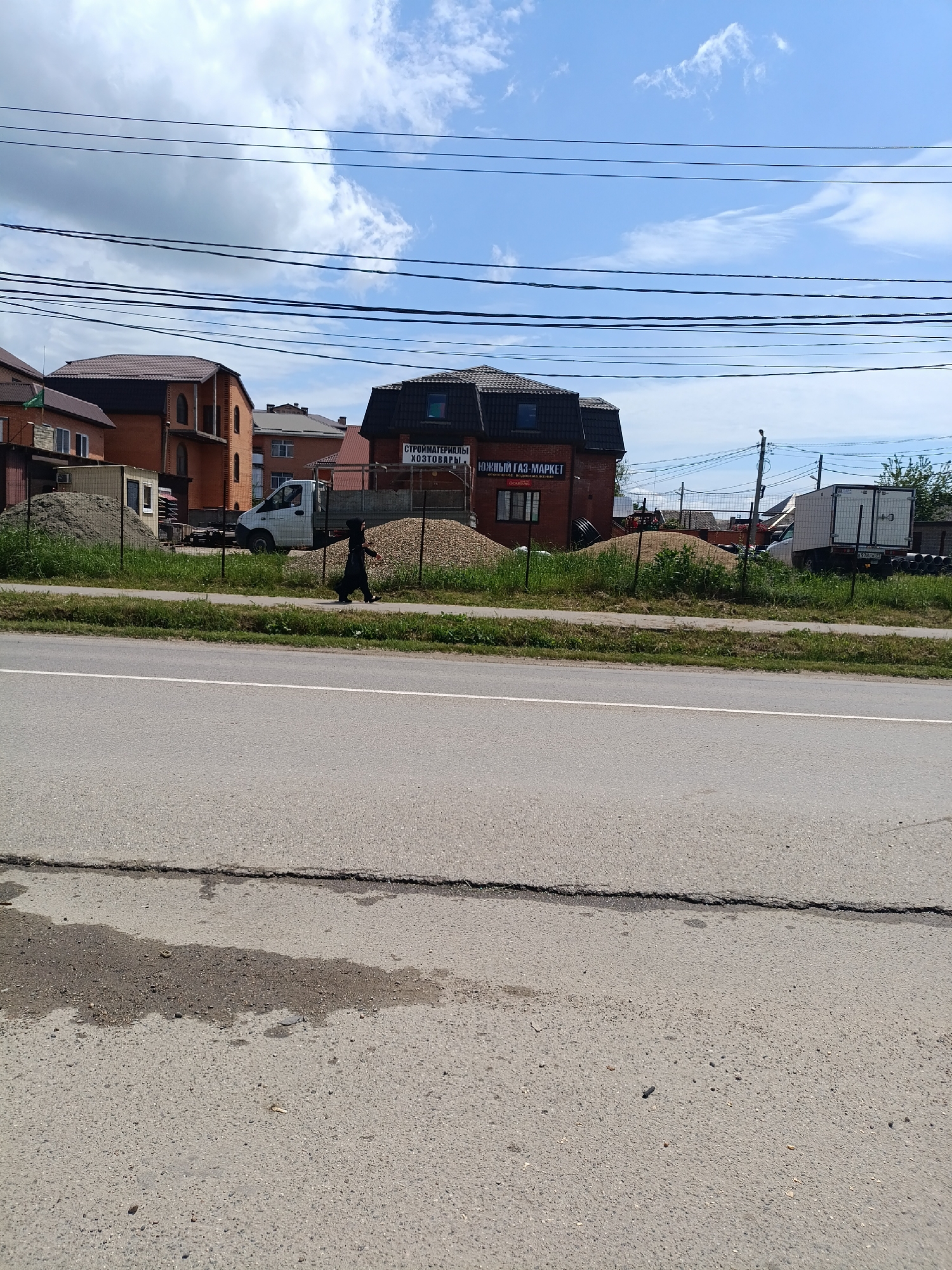 The image captures a street scene in Yablonovskiy, Russia, featuring a paved road in the foreground with a long crack running across its surface. A dark, irregular stain is visible on the asphalt. Beyond the road, there's a narrow grassy verge with a low chain-link fence. Behind the fence are piles of gravel and sand, construction materials seemingly related to a building supply store, "Южный газ-маркет" (Southern Gas Market), visible in the background. The store is a two-story brick building with a dark roof. Adjacent to the store, there is a commercial van and other vehicles. A person dressed in dark clothing is walking along the sidewalk, between the gravel piles and the van. Other buildings, primarily residential houses with brick facades and brown roofs, are partially visible in the background. Overhead, several black power lines run across the frame against a sky with scattered clouds.