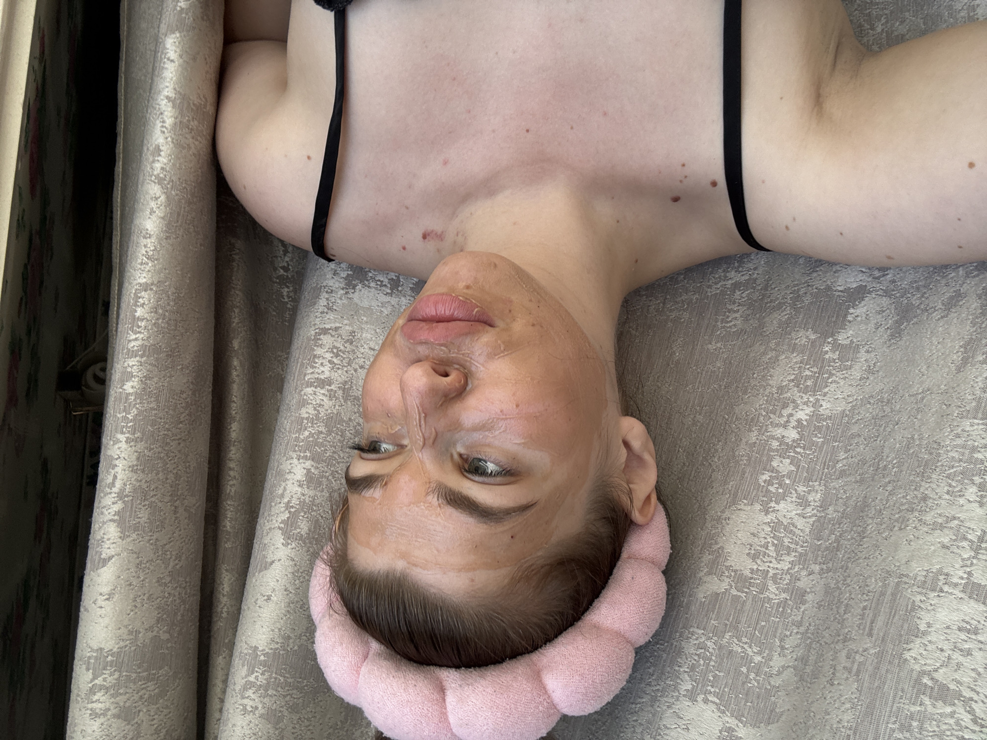 A person, possibly female, is lying upside down with a pink headband, a facial mask and a black tank top. The background consists of patterned gray fabric or curtains. The location in the city of Мещёры, Russia is not discernible from the image.