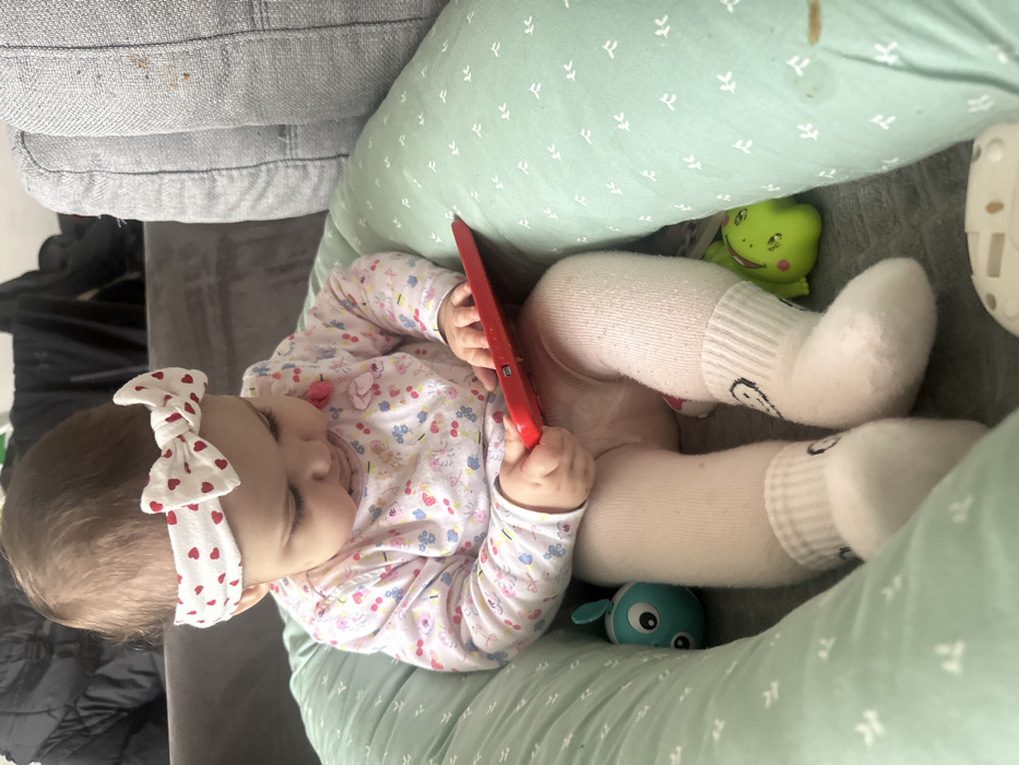 In an indoor, domestic setting within Strasbourg, France, an infant is seated upright in a mint green padded support pillow adorned with a light geometric pattern. The infant wears a white headband with red hearts, a long-sleeved light-colored top featuring a small floral or fruit pattern, light-colored tights or leggings, and white socks with a black smiley face design on the cuffs. The infant is holding a red, rectangular device, possibly a tablet or toy, with both hands and is looking down intently at it. To the infant's left, a teal-colored toy with large white eyes is visible within the support pillow. To the infant's right, a green toy, appearing to be a frog or similar creature, rests. In the background, behind the infant, is a grey upholstered sofa, which shows some visible brown staining on its surface. A black garment or object is partially visible on another grey surface further in the background. A white object is partially visible on the floor or surface in front of the infant's feet.