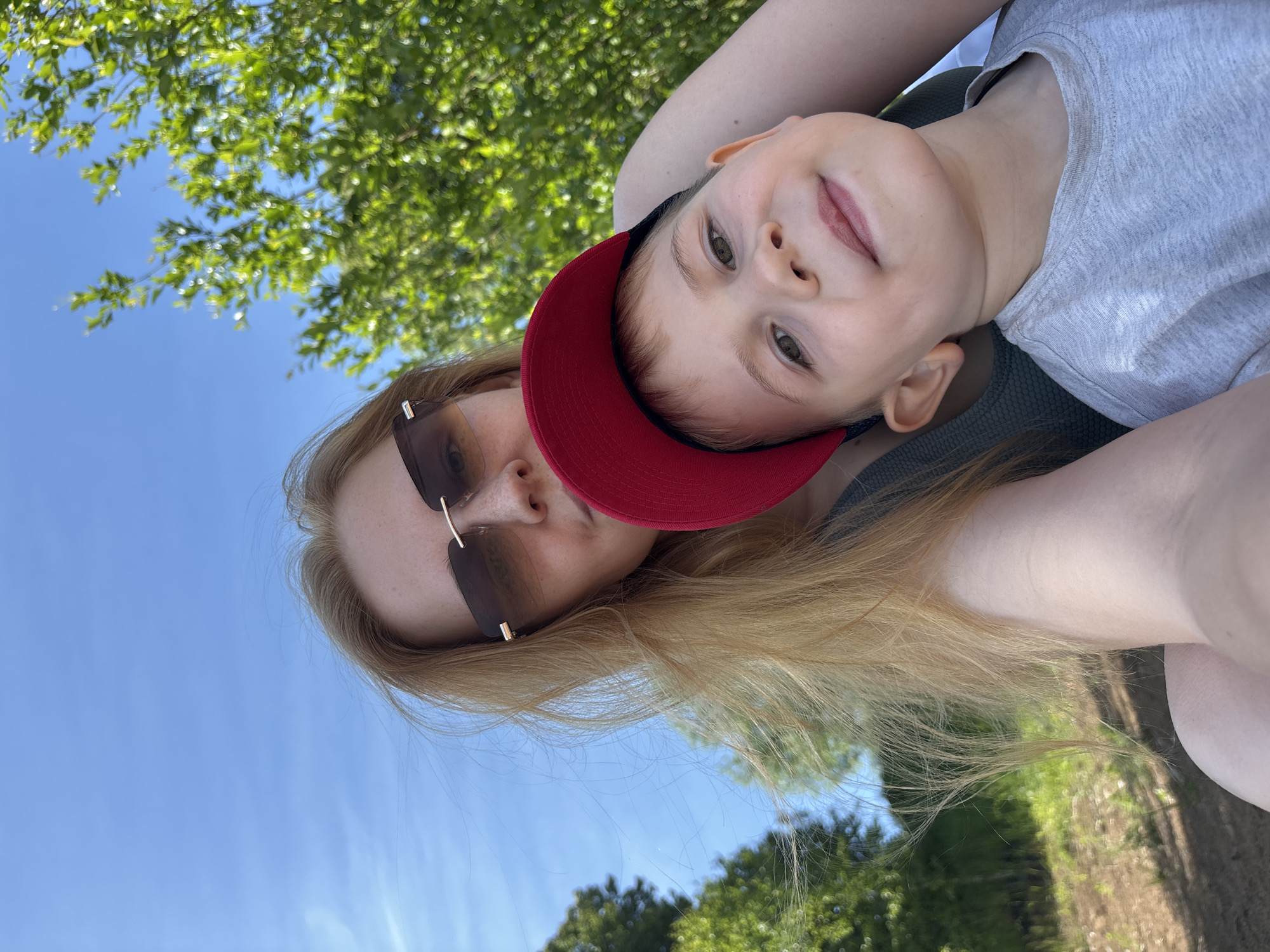 A blonde woman with sunglasses and a child wearing a red cap are posing for a selfie outdoors. The woman's hair is blowing in the wind, and she is wearing a green top. The child is wearing a gray t-shirt and has a bright smile. They are surrounded by lush green trees and a bright blue sky, suggesting they are in a park or a natural setting within the city of Unknown. The lighting indicates it is daytime.