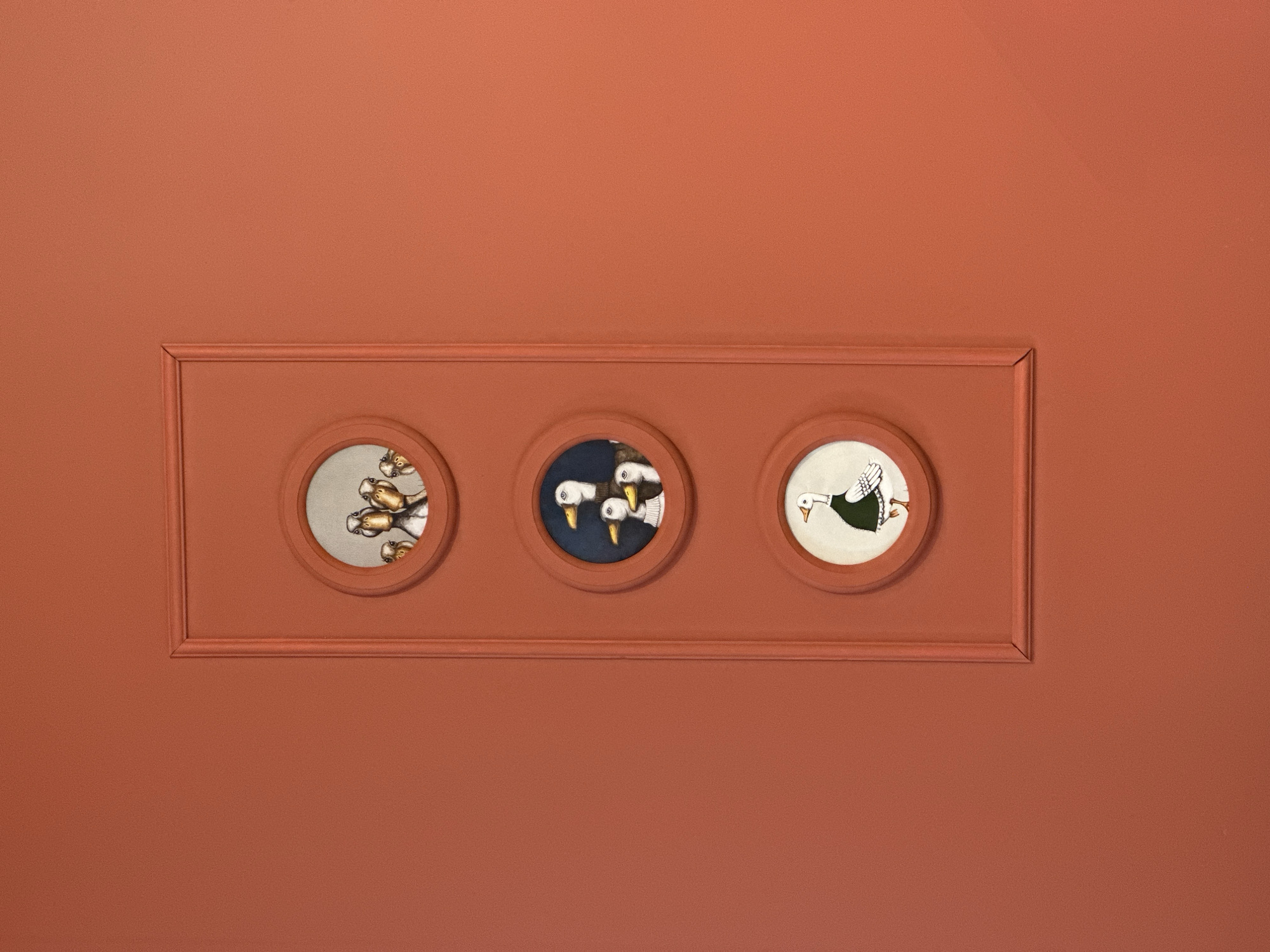 The image features a decorative panel with three circular insets displaying ornithological themed artwork. The panel and the wall behind it are painted in a uniform terracotta color. The left inset depicts several geese. The central inset shows three geese heads against a dark background. The right inset displays a goose with an unusual outfit. The scene has a decorative and artistic atmosphere. The location is assumed to be somewhere in Chaikovsky, Russia.