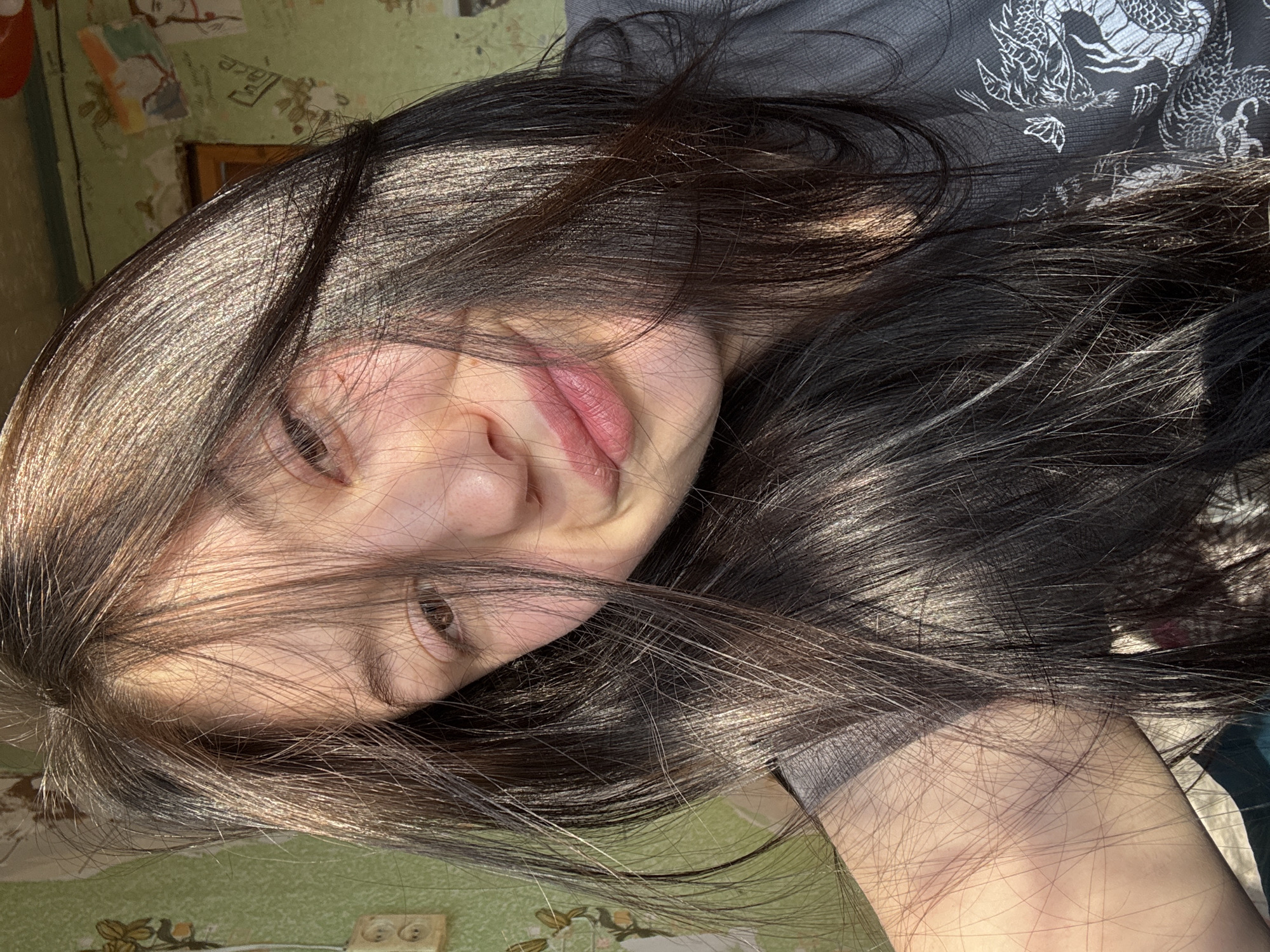 A person is lying on their side, with their face and dark hair angled towards the camera. The person appears to be wearing a dark t-shirt with a white dragon design. The background includes a textured green wall with visible electrical outlets. The location is indicated as Фоки, Russia.